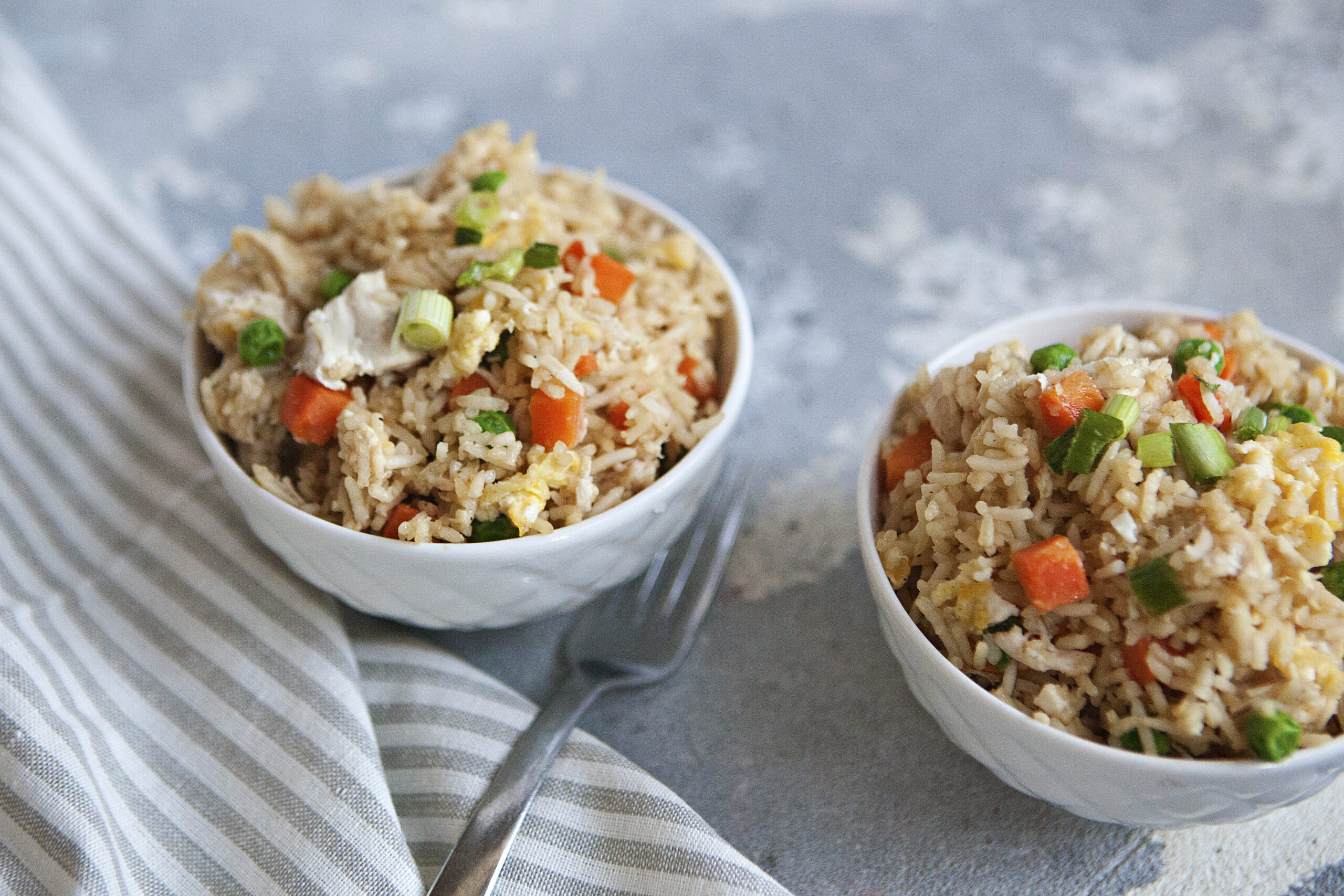 A bowl of chicken fried rice with egg, peas, carrots, and green onion on a gray background.