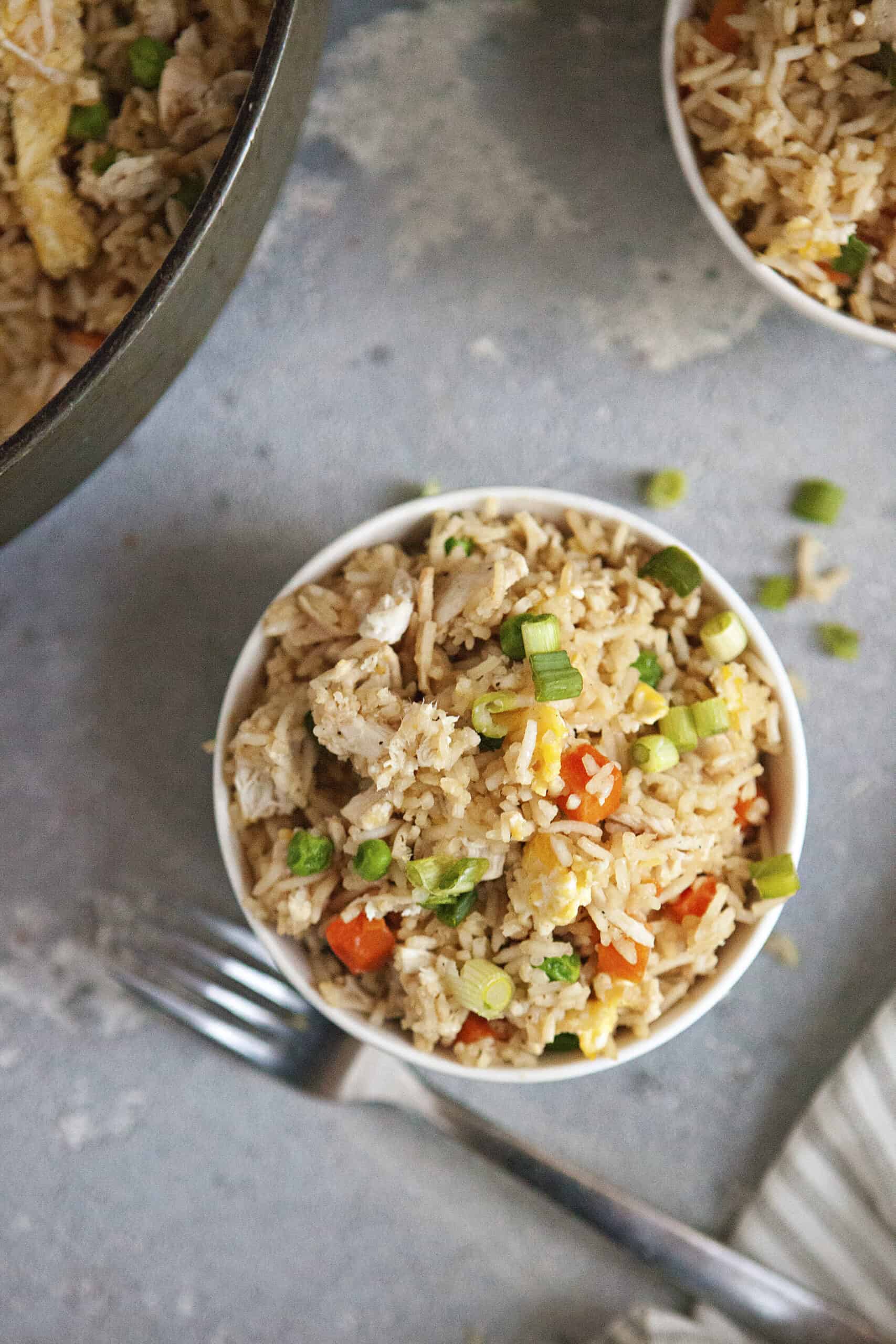 A bowl of chicken fried rice with egg, peas, carrots, and green onion on a gray background with a fork.