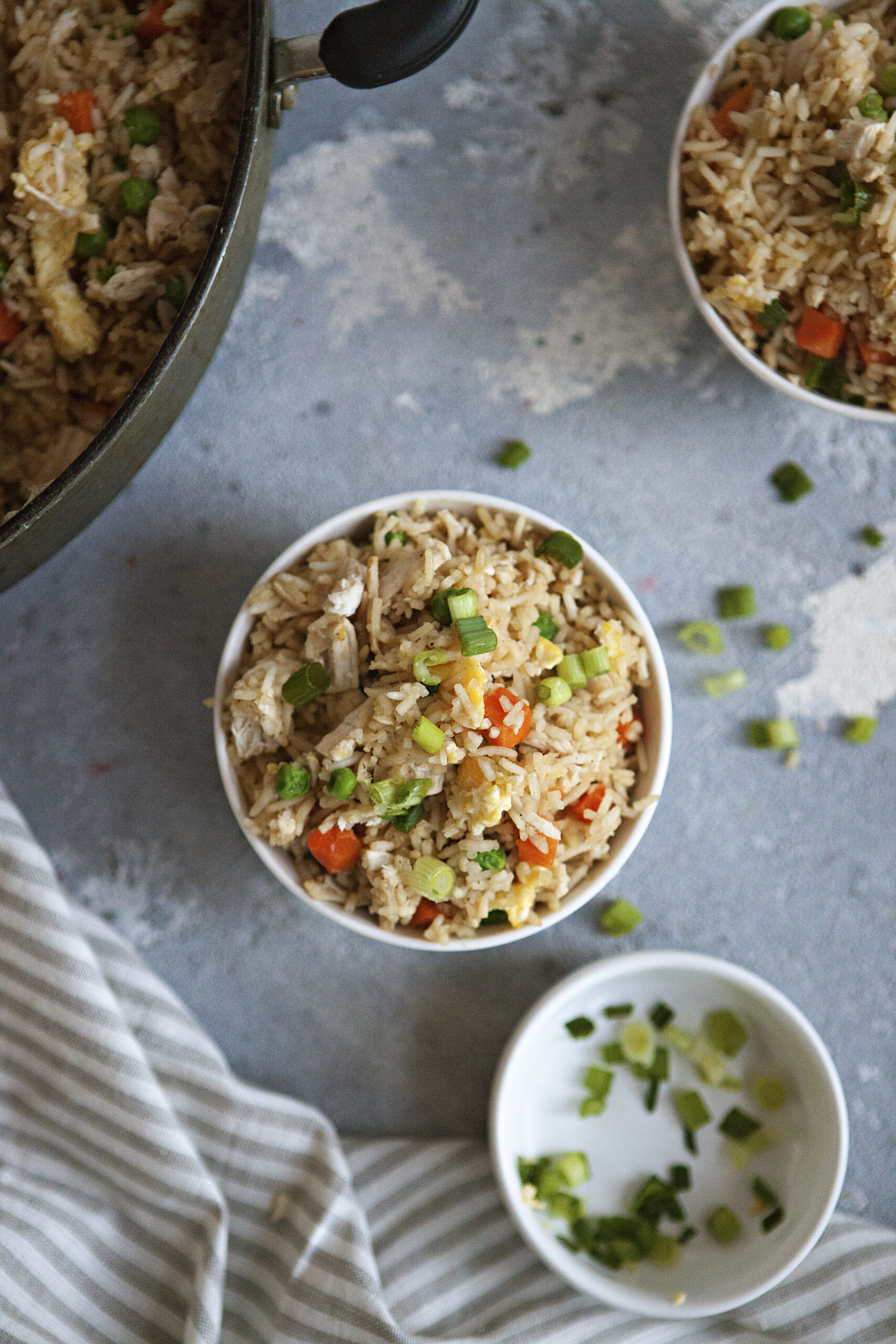 A bowl of chicken fried rice with egg, peas, carrots, and green onion on a gray background.