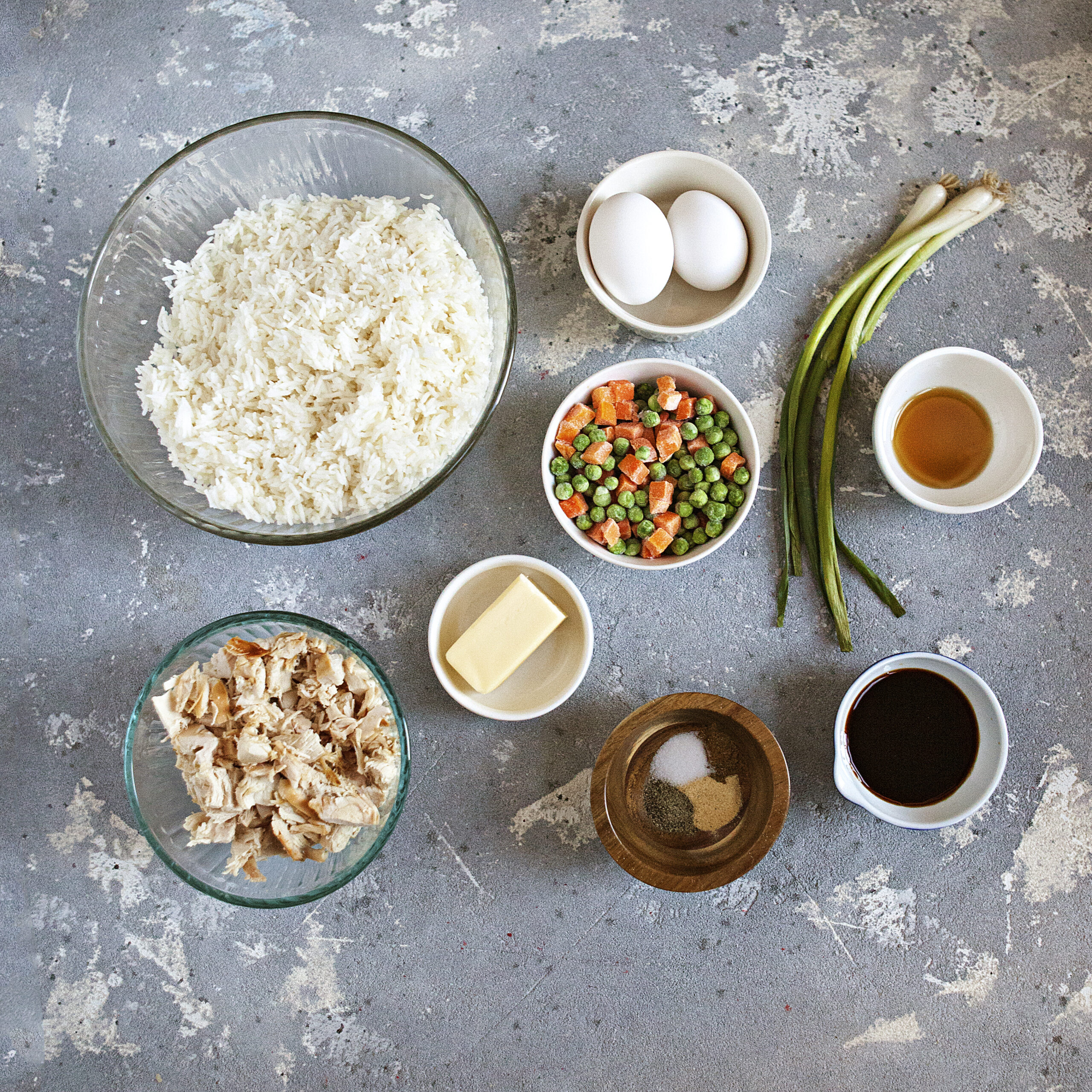 Ingredients for chicken fried rice:  Leftover white rice, diced chicken, butter, frozen peas and carrots, fresh green onions, 2 eggs, soy sauce, sesame oil, salt, pepper, garlic powder, and Chinese 5-spice powder, on a gray background.