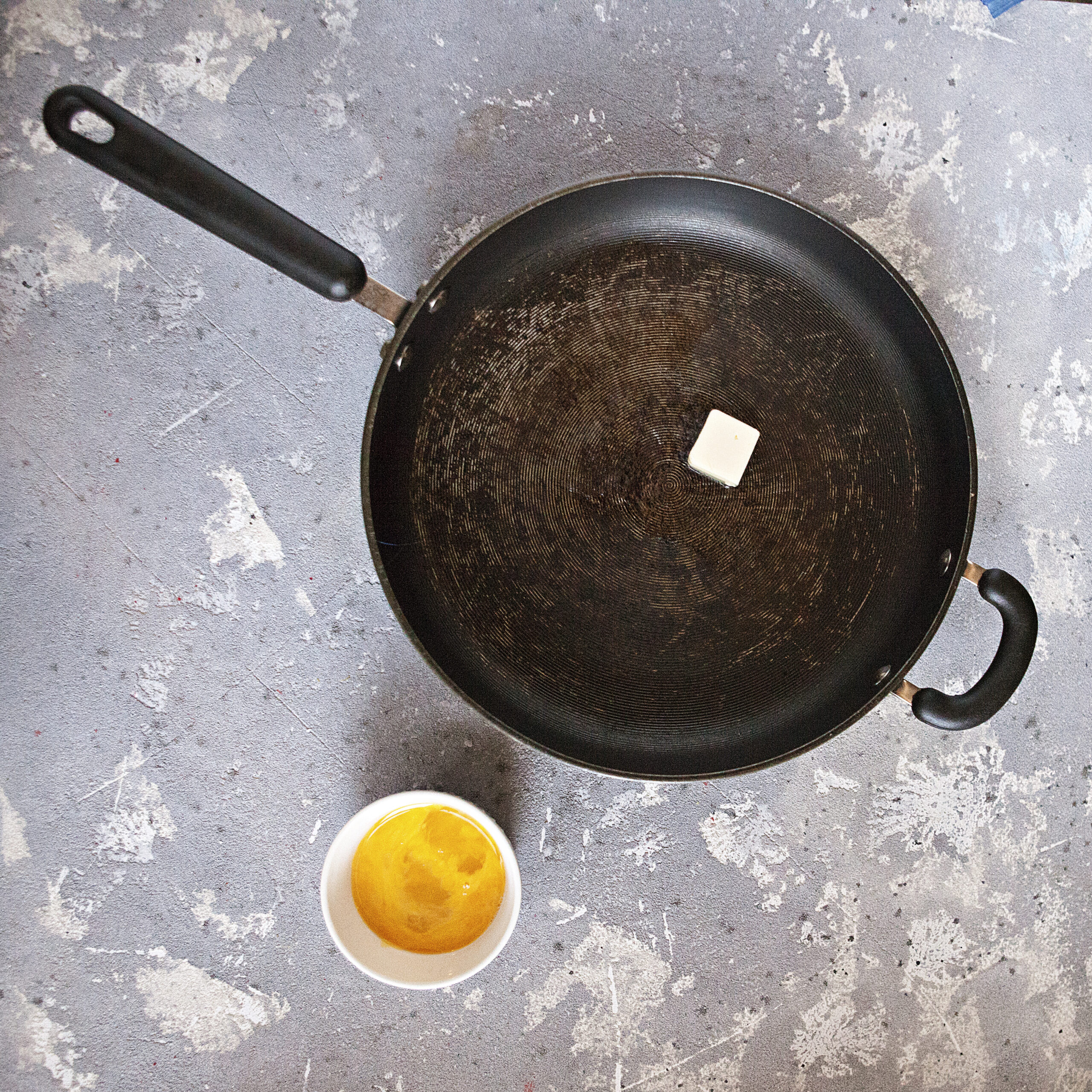 A black saucepan with a tablespoon of butter next to a small dish with 2 whisked eggs on a gray background.