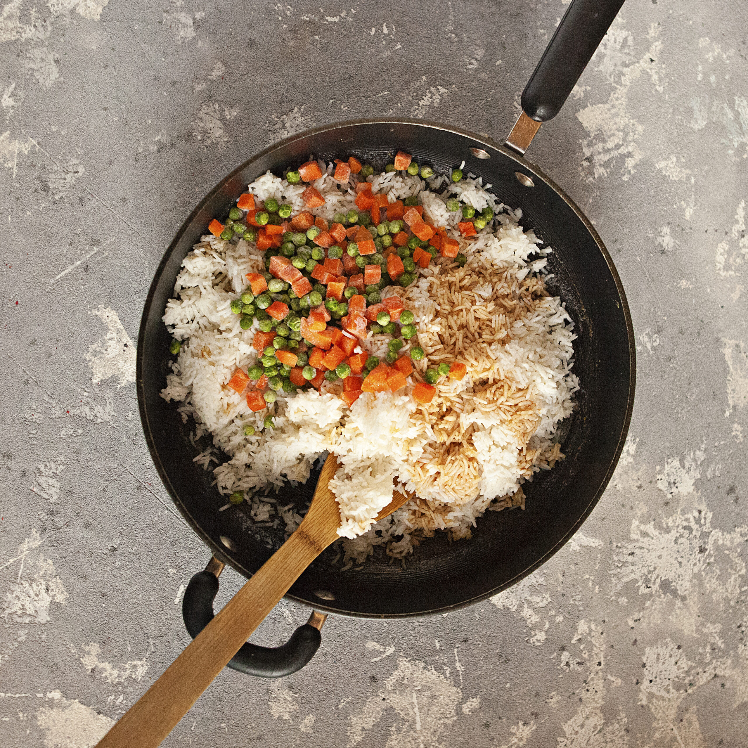 A black skillet containing white rice, frozen peas and carrots, spices, and soy sauce with a wooden spoon.