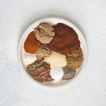 Spices for taco seasoning mix on a plate: Chili powder, brown sugar, smoked paprika, cumin, garlic powder, coriander, onion powder, oregano, cocoa powder, salt, pepper, on a white background.