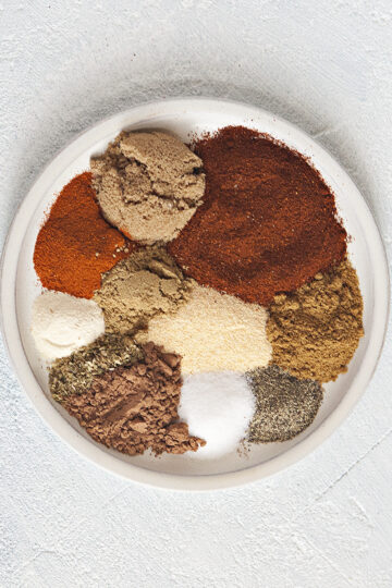 Spices for taco seasoning mix on a plate: Chili powder, brown sugar, smoked paprika, cumin, garlic powder, coriander, onion powder, oregano, cocoa powder, salt, pepper, on a white background.