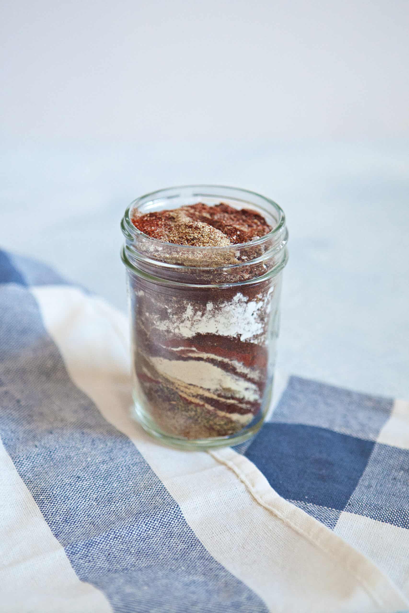 Ingredients for homemade taco seasoning mix in a mason jar on a blue and white kitchen towel.