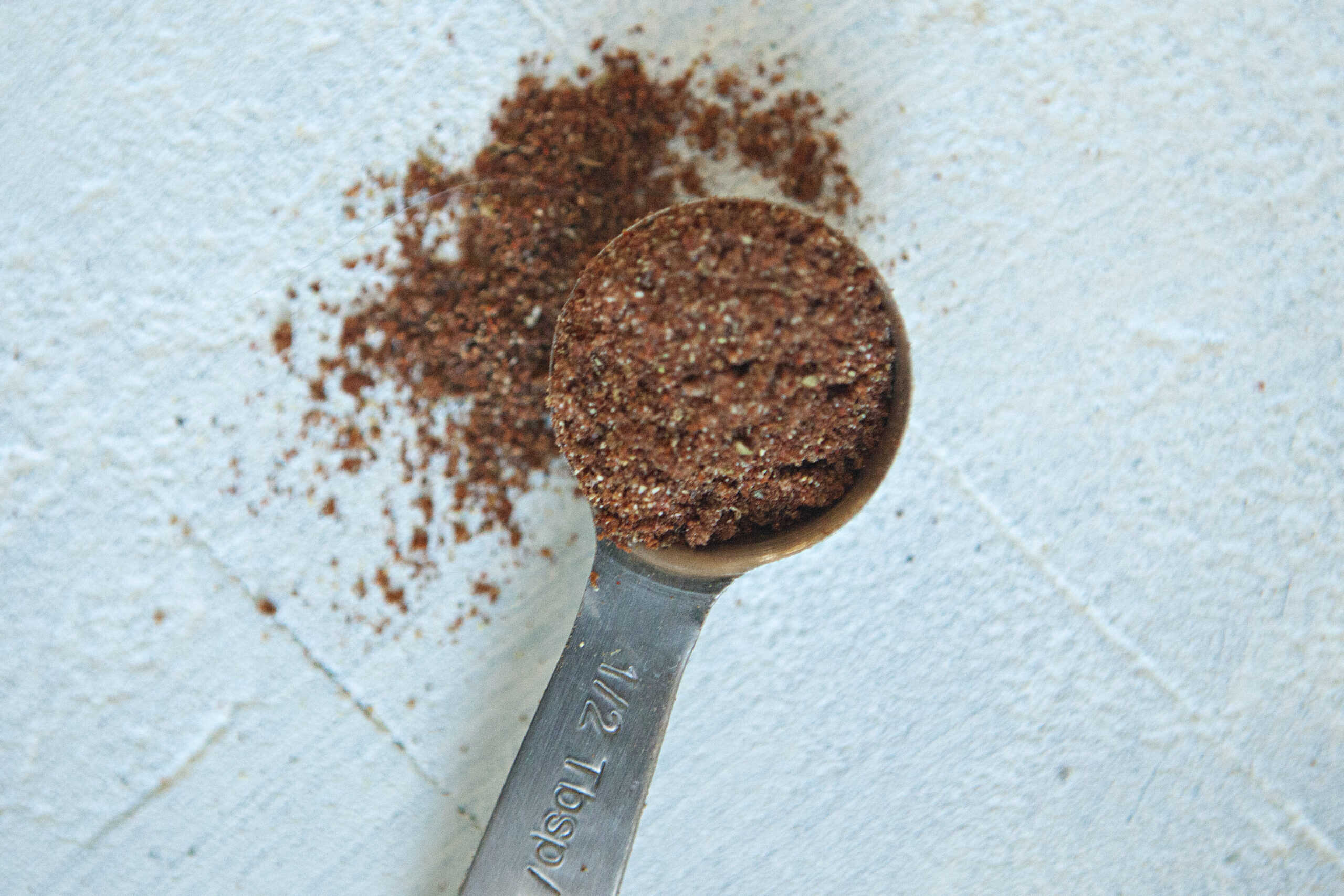 Dark red-orange taco seasoning mix in a metal measuring spoon, spilling over onto a white background.