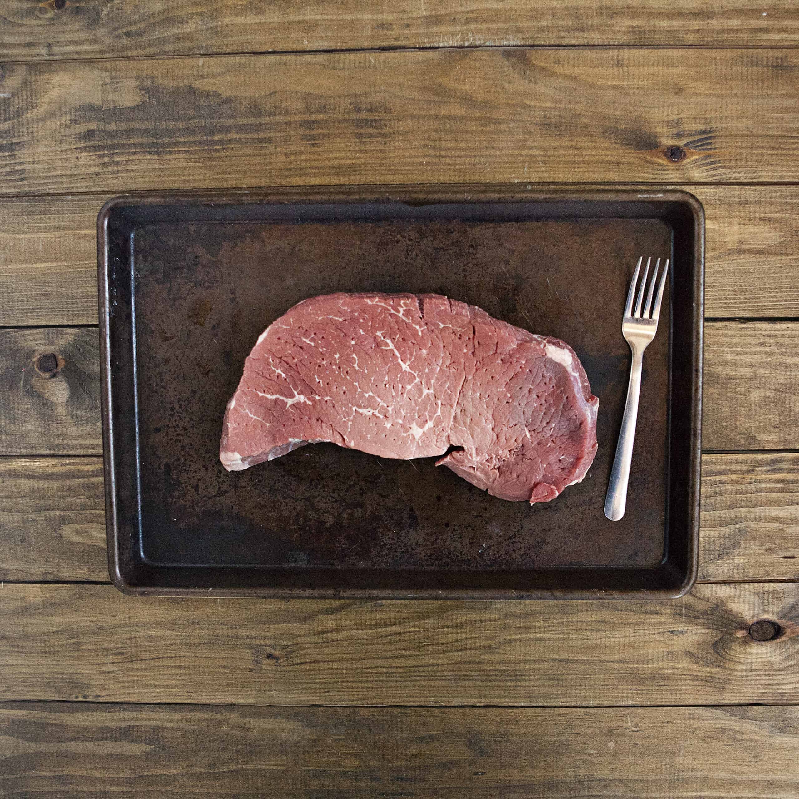 A raw London broil steak that has been tenderized with a fork on a baking pan on a wooden table.