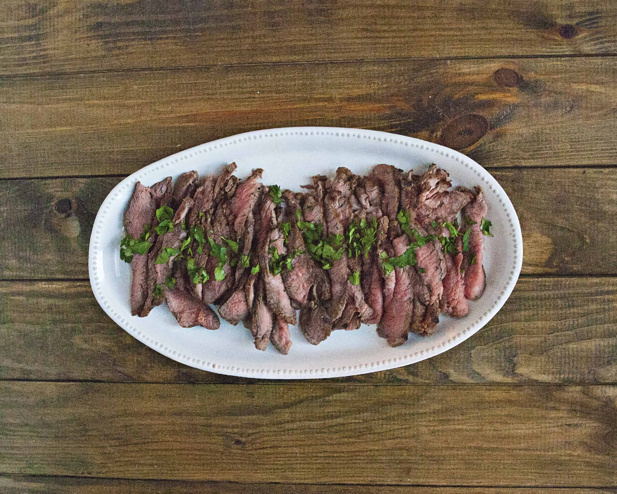 Sliced medium-rare London broil steak topped with chopped parsley on a white serving platter on a brown wooden table.