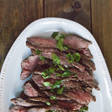 Sliced London broil steak topped with chopped parsley on a white serving platter on a brown wooden table.