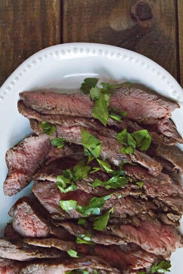 Sliced London broil steak topped with chopped parsley on a white serving platter on a brown wooden table.