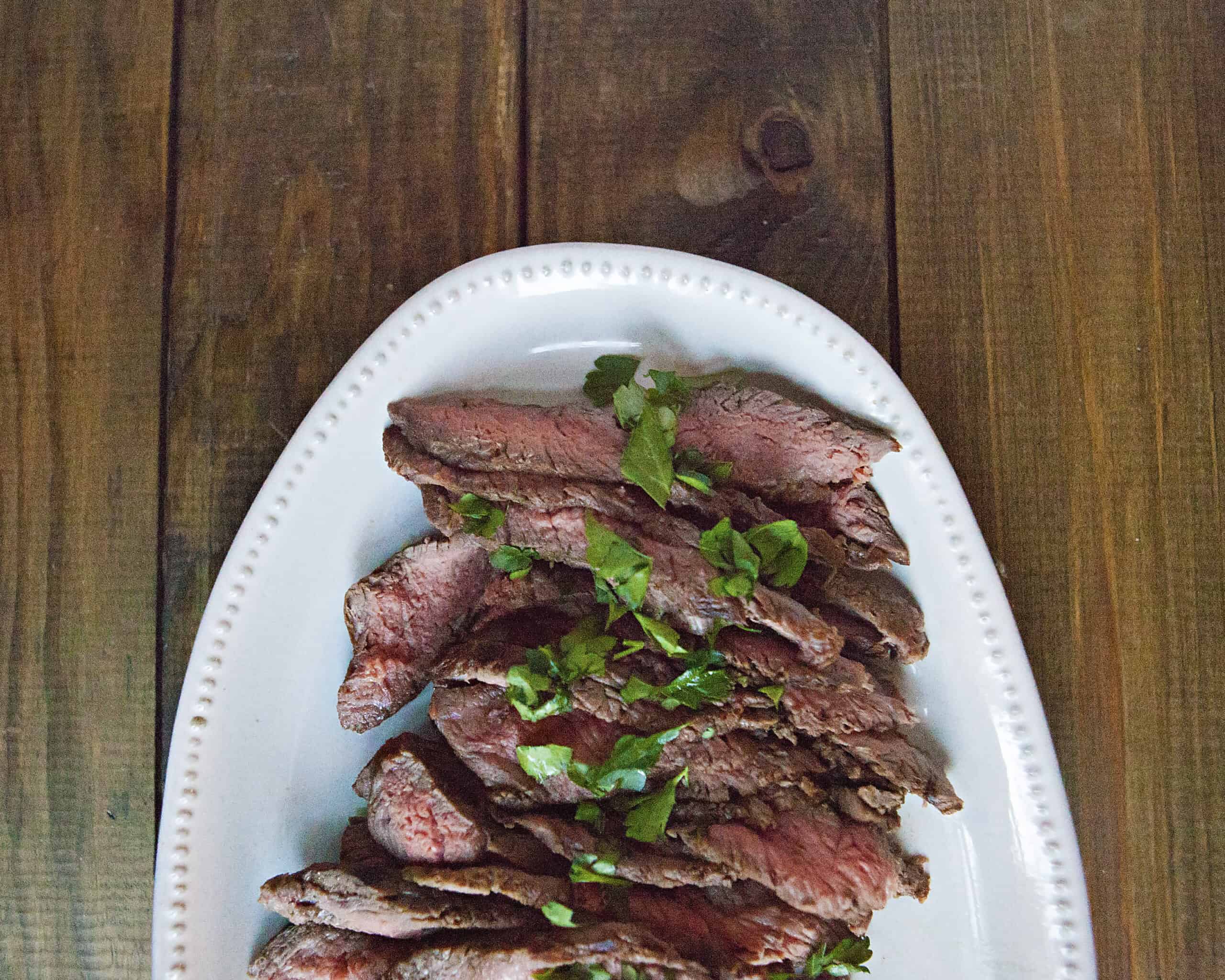 Sliced London broil steak topped with chopped parsley on a white serving platter on a brown wooden table.