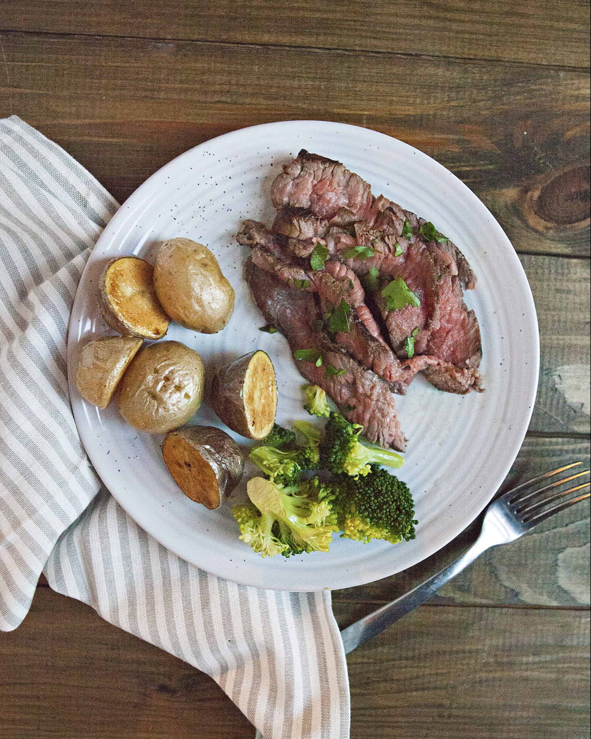 A white plate containing London broil steak with chopped parsley, roasted baby potatoes, and steamed broccoli on a brown wooden table alongside a fork and a gray-and-white striped napkin.