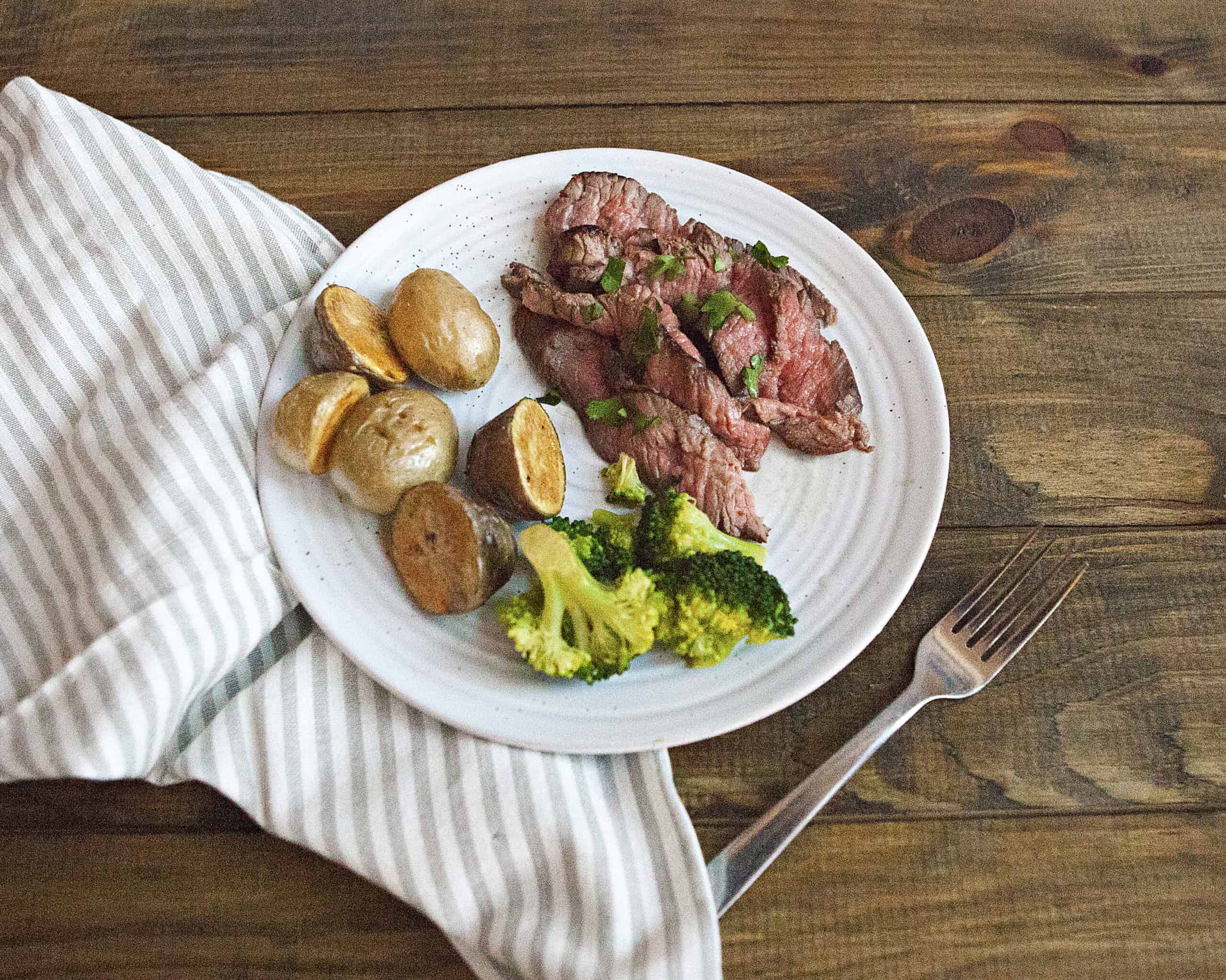 A white plate containing London broil steak with chopped parsley, roasted baby potatoes, and steamed broccoli on a brown wooden table alongside a fork and a gray-and-white striped napkin.