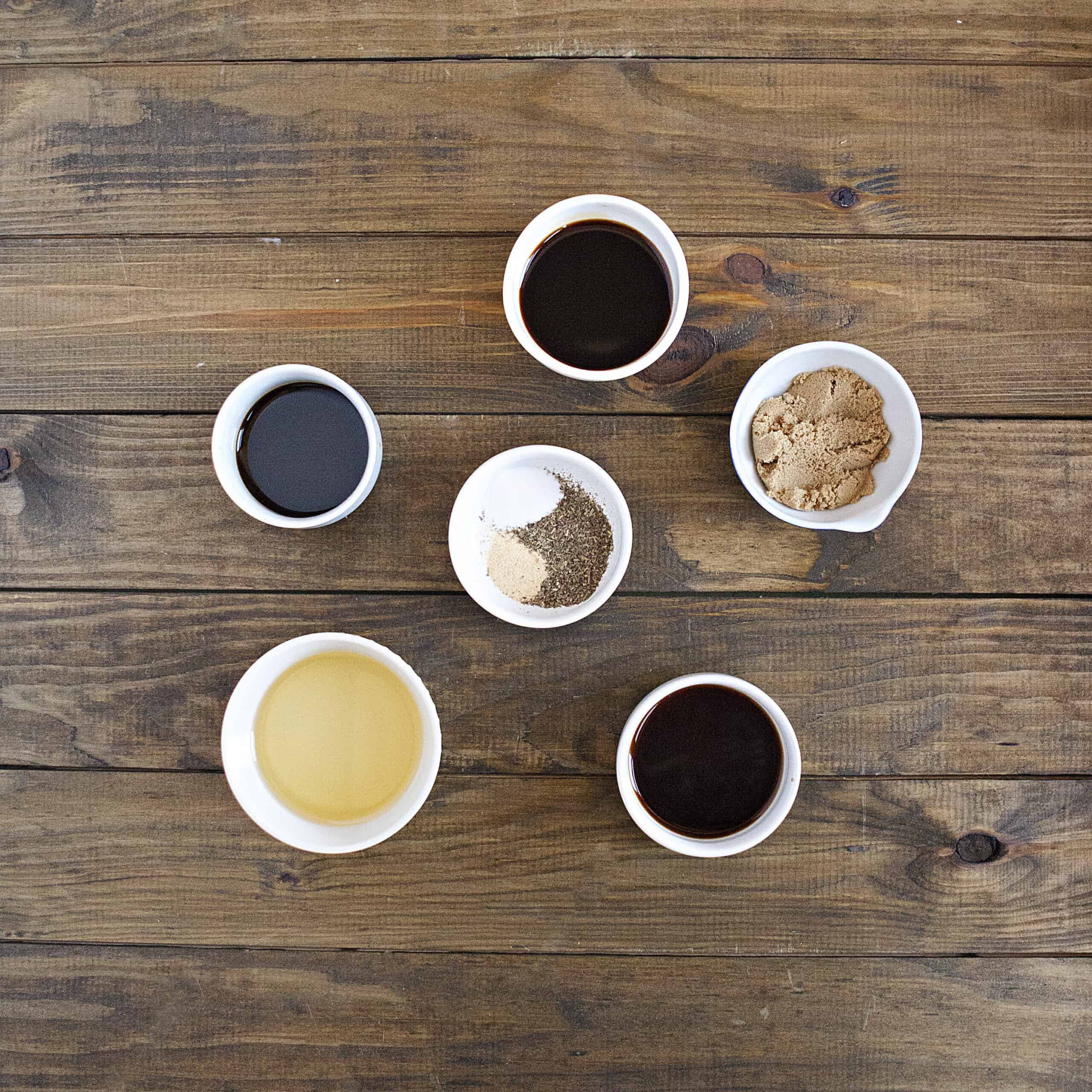 Ingredients for balsamic marinade for London broil steak:  Soy sauce, balsamic vinegar, Worcestershire sauce, olive oil, brown sugar, salt, garlic powder, and Italian seasoning, on a brown wooden table.