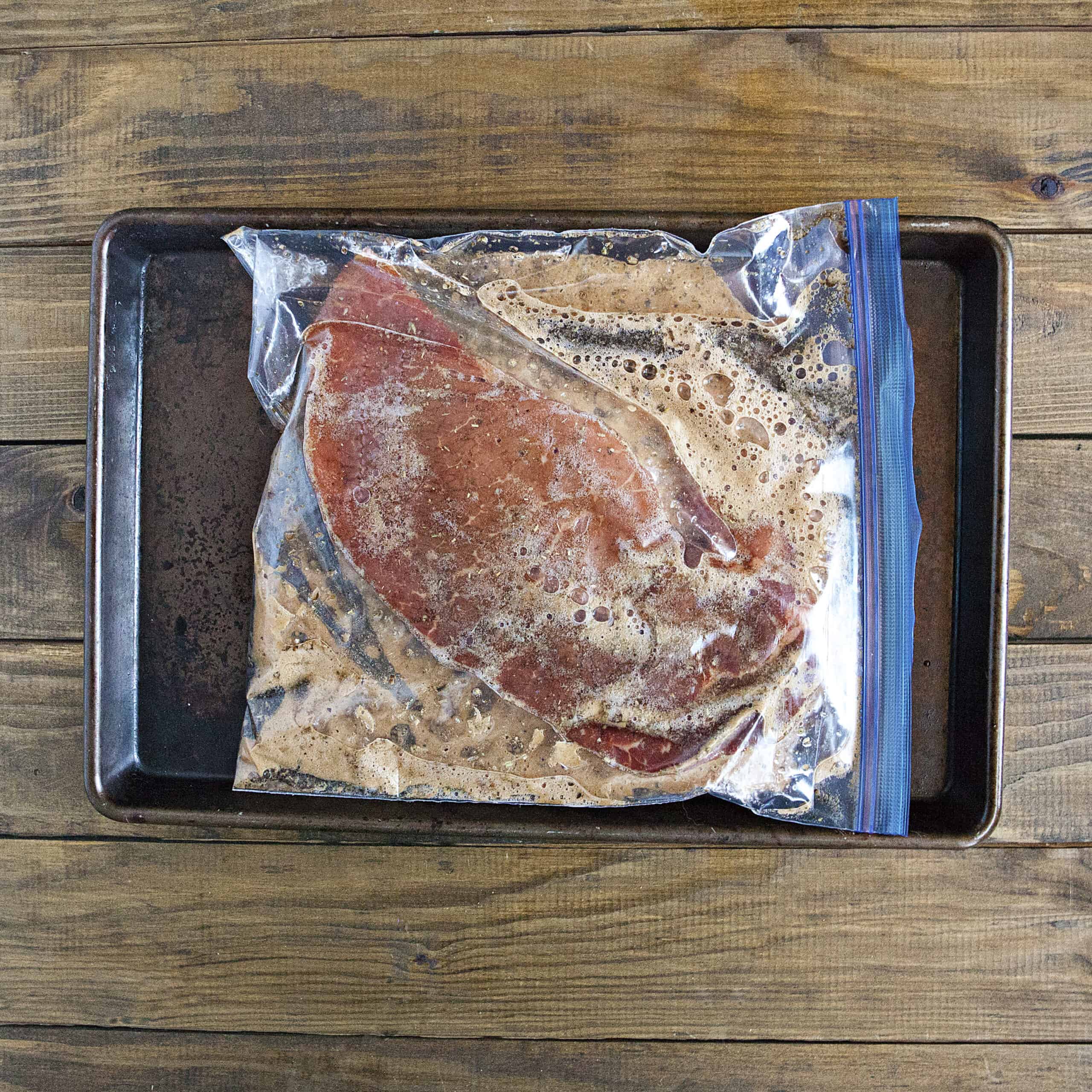 London broil steak in a balsamic marinade in a zip-top plastic bag on a baking sheet on a brown wooden table.