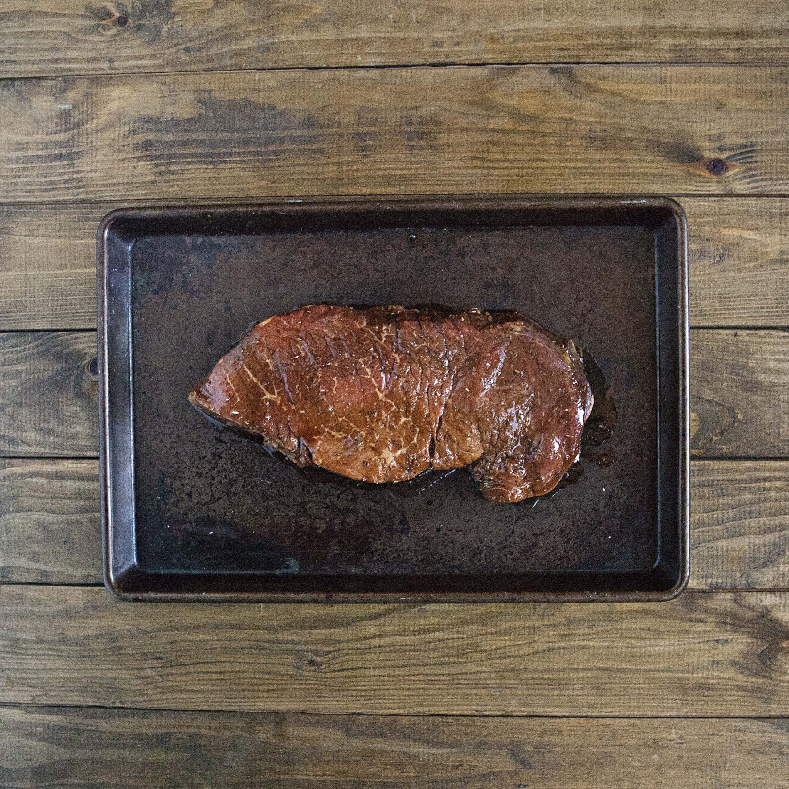 A juicy raw marinated London broil steak on a baking pan on a brown wooden table.