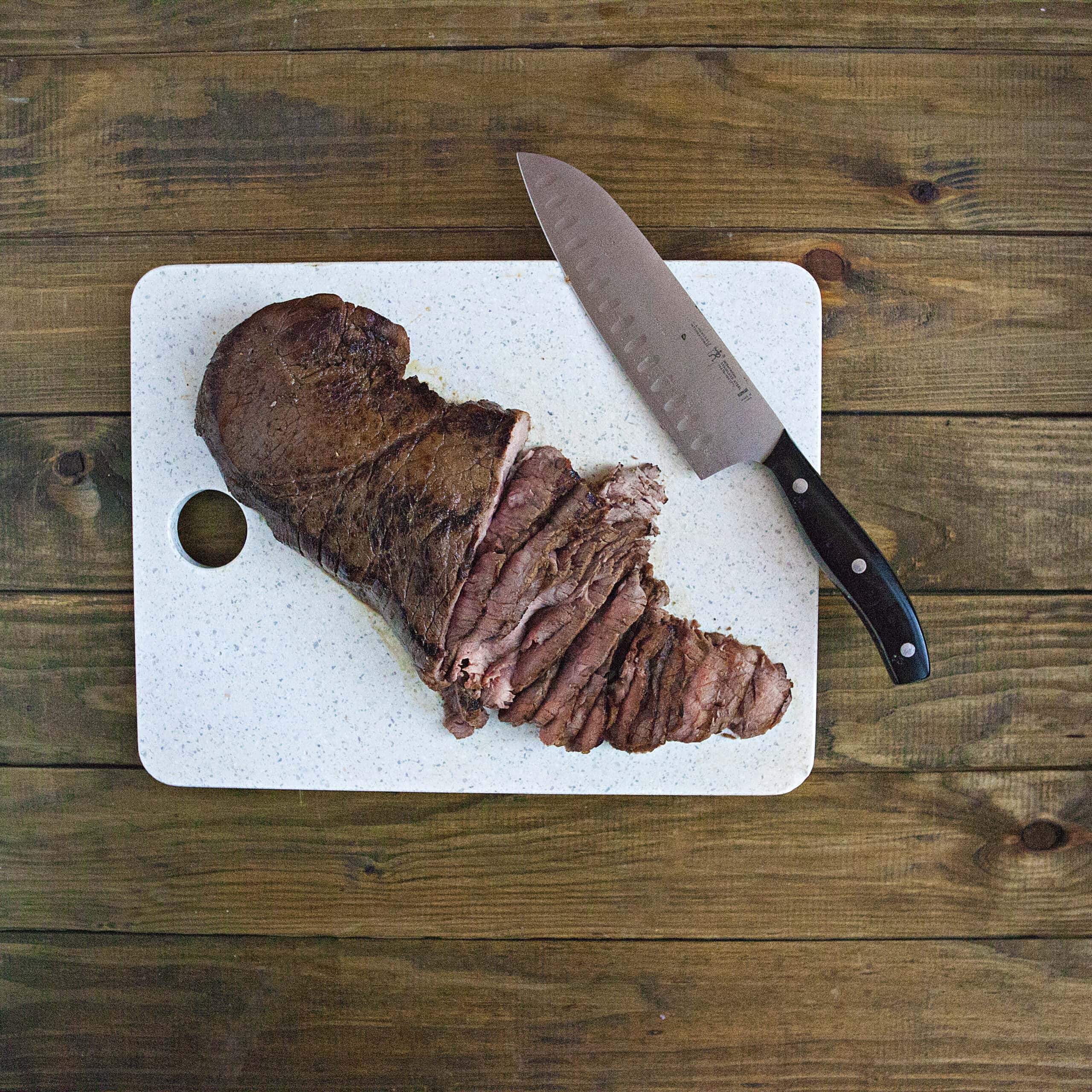 Partially sliced London broil steak on a white cutting board with a knife on a brown wooden table.