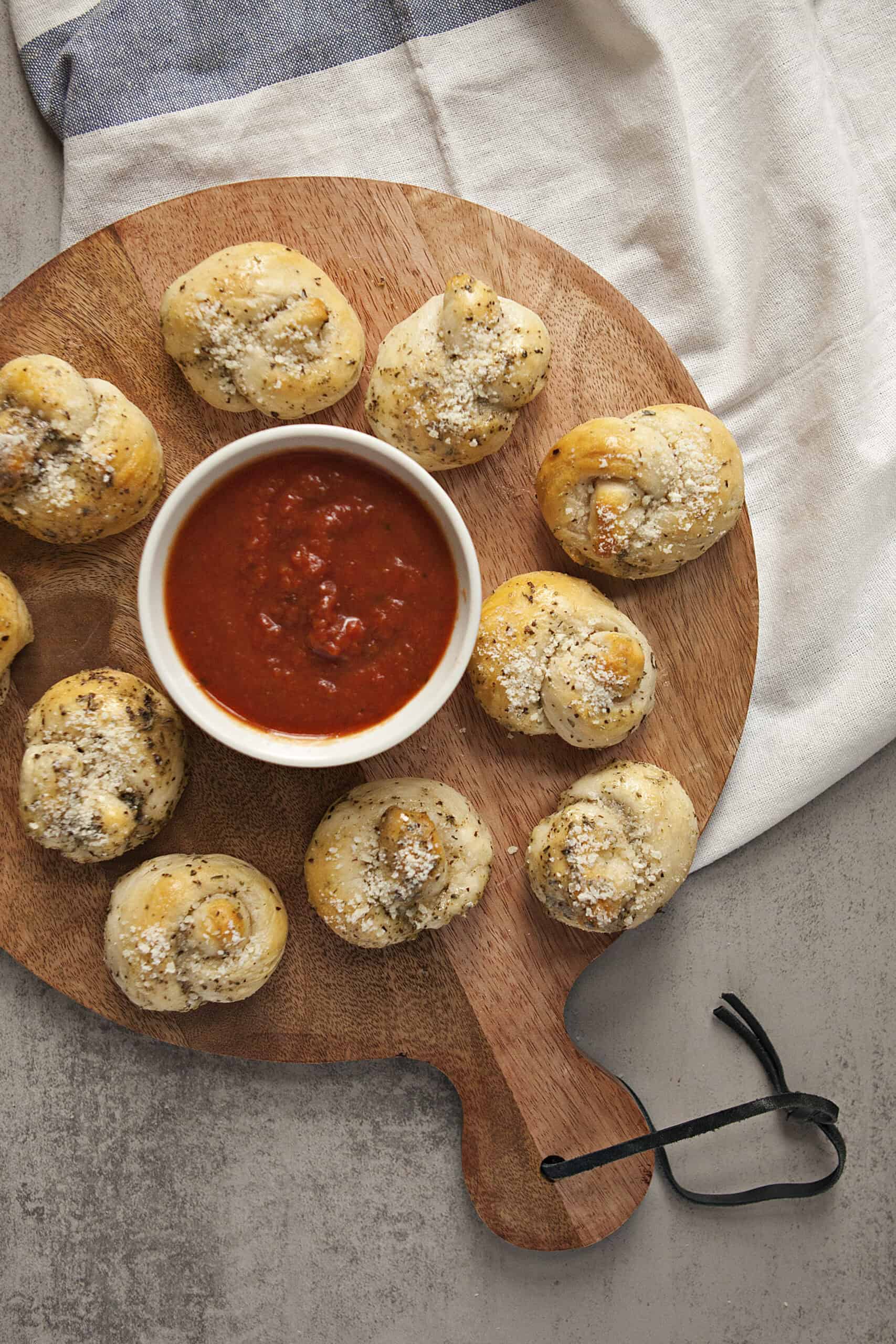 A round serving board holding buttery Parmesan garlic knots and a ramekin containing marinara dipping sauce.