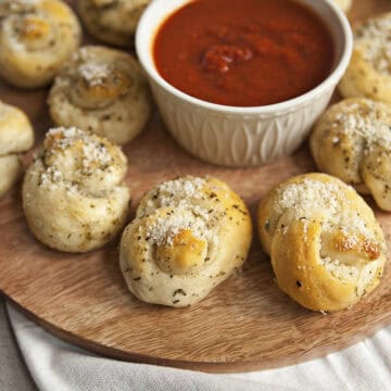 Buttery Parmesan garlic knots on a round serving tray with a white ramekin filled with marinara sauce.