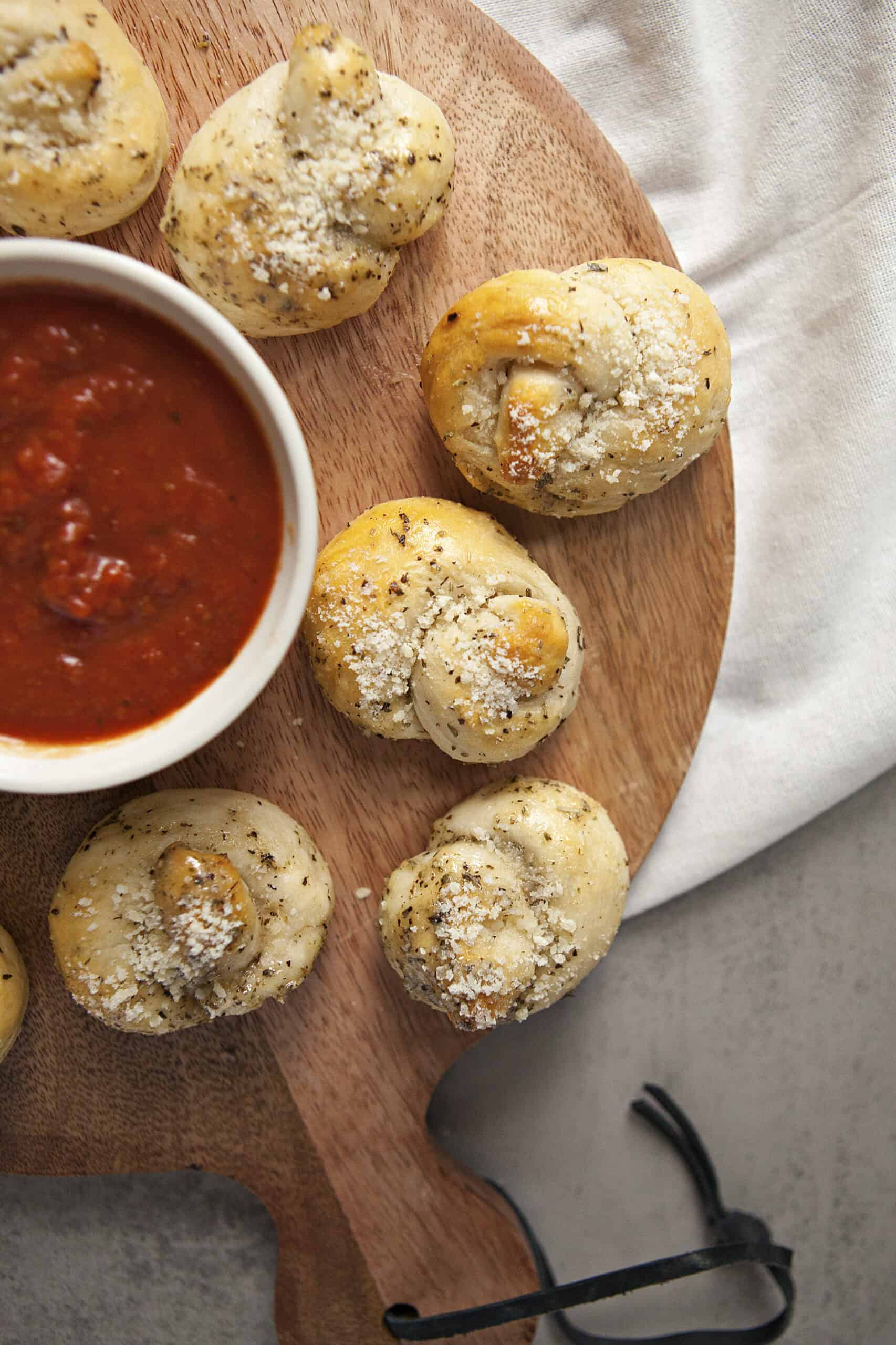 Buttery Parmesan garlic knots on a wooden cutting board next to a ramekin of marinara sauce.