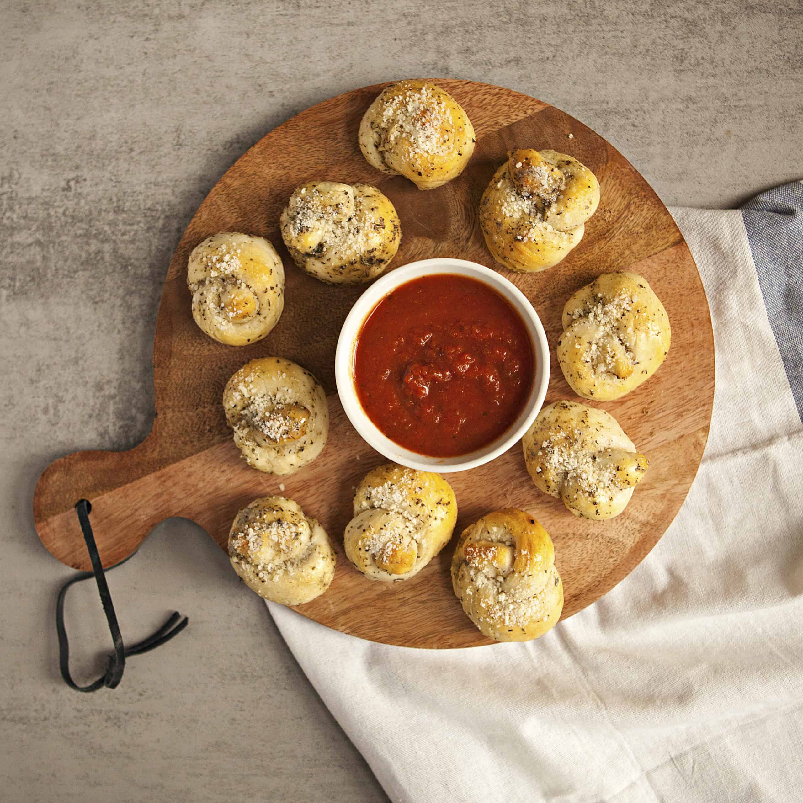 A round serving board holding buttery Parmesan garlic knots and a ramekin containing marinara dipping sauce.