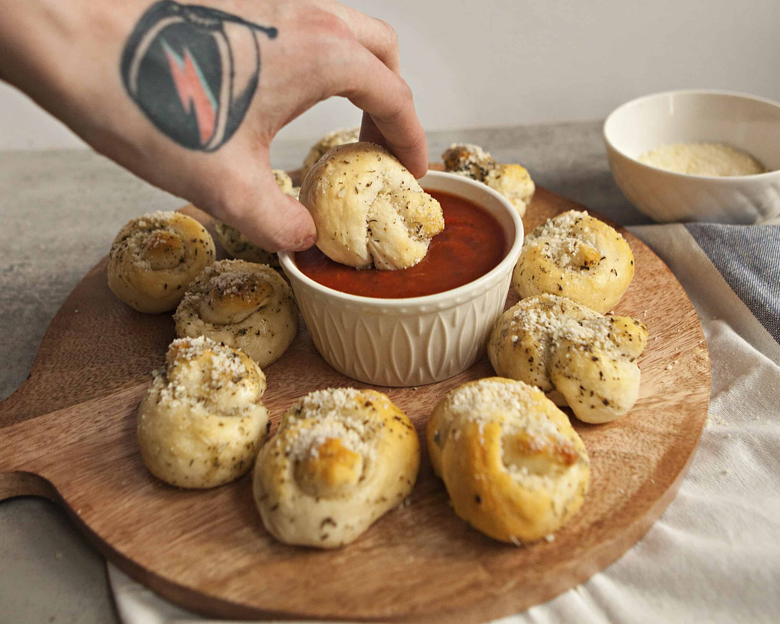 A tattooed hand holding a buttery Parmesan garlic knot being dipped into marinara sauce, above a wooden serving platter containing more garlic knots.