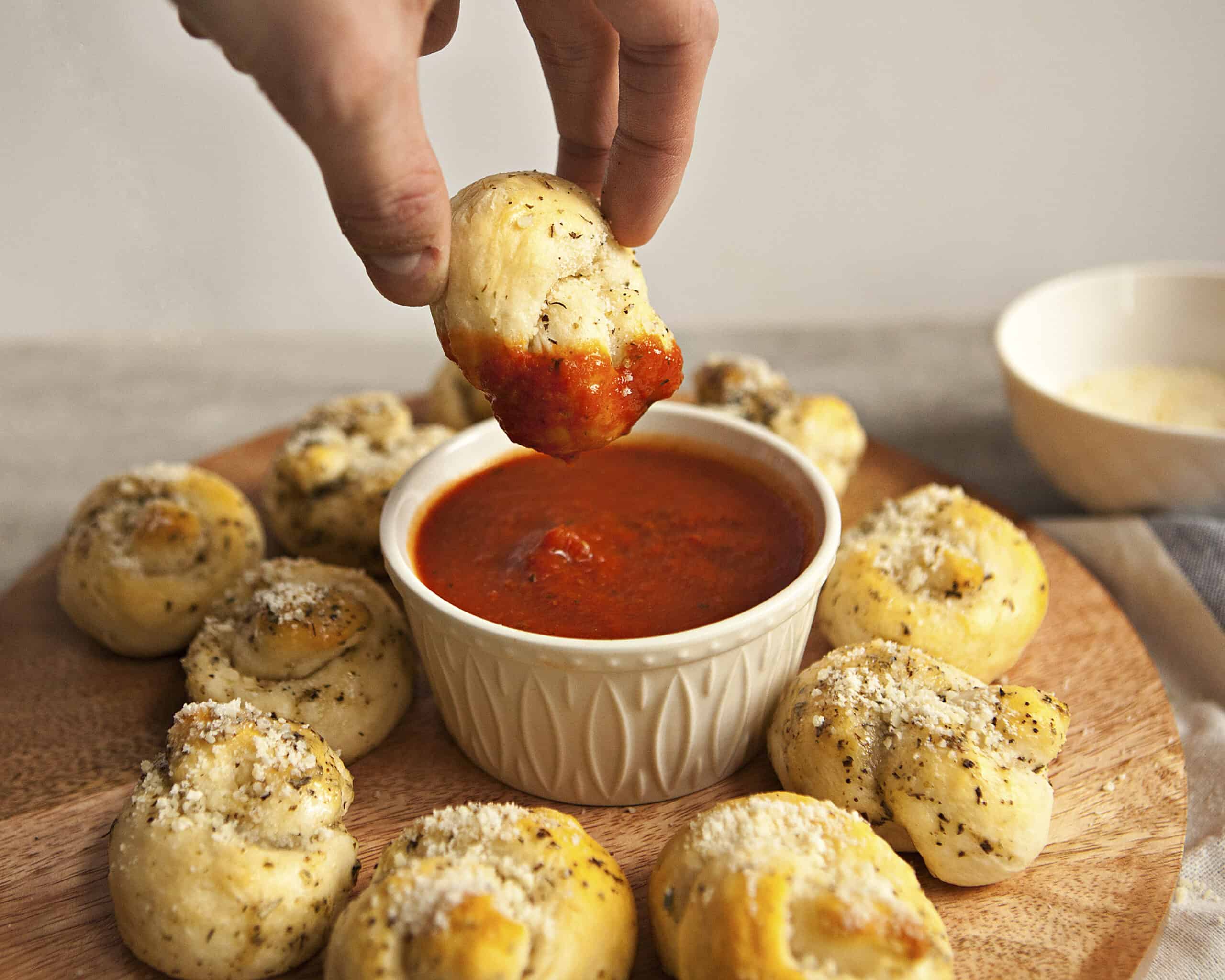 A hand holding a garlic knot partially coated in marinara sauce, above a ramekin containing more marinara sauce and a serving board with more garlic knots.