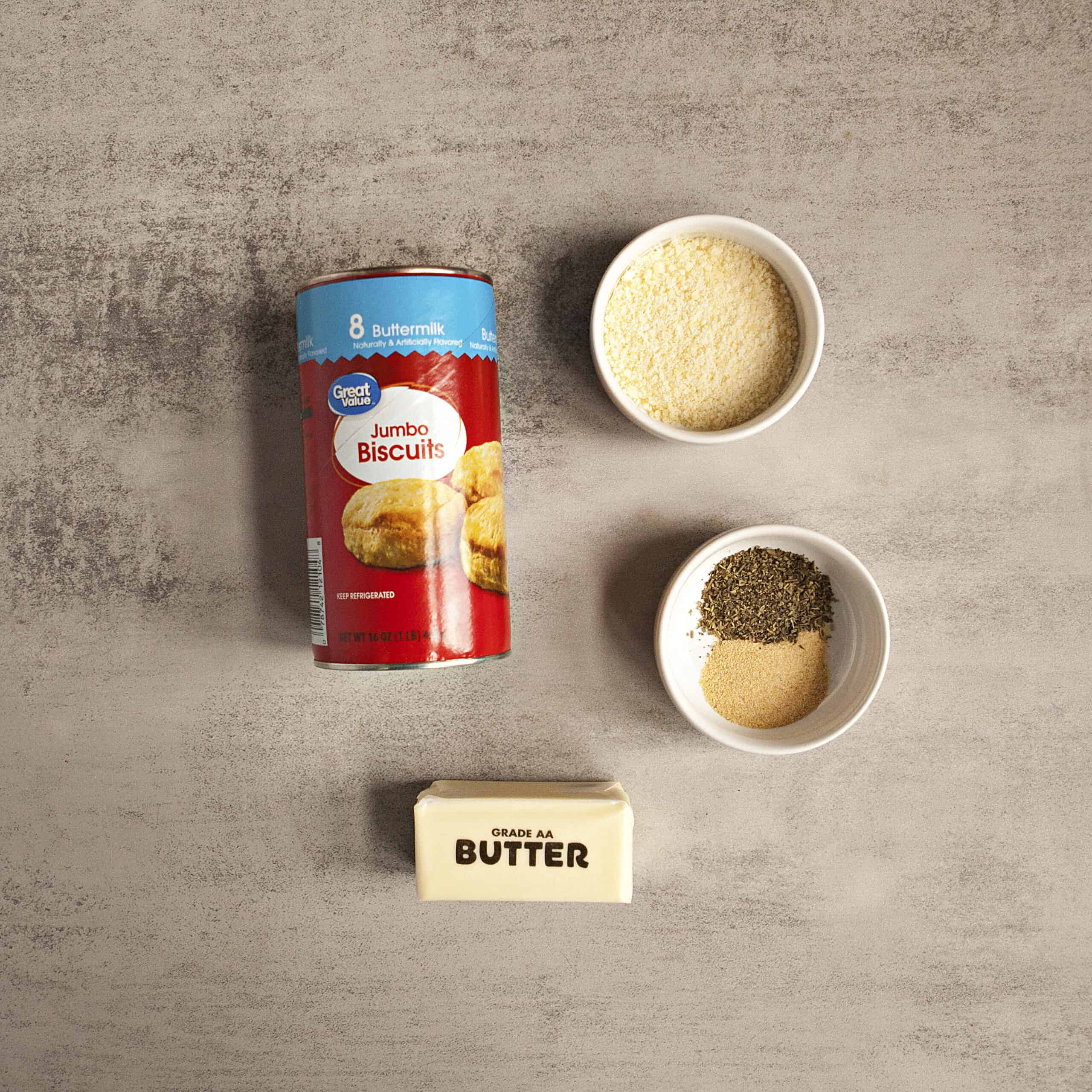 Ingredients for garlic knots:  store-bought biscuit dough, grated Parmesan cheese, butter, garlic powder, and Italian seasoning, on a gray backdrop.