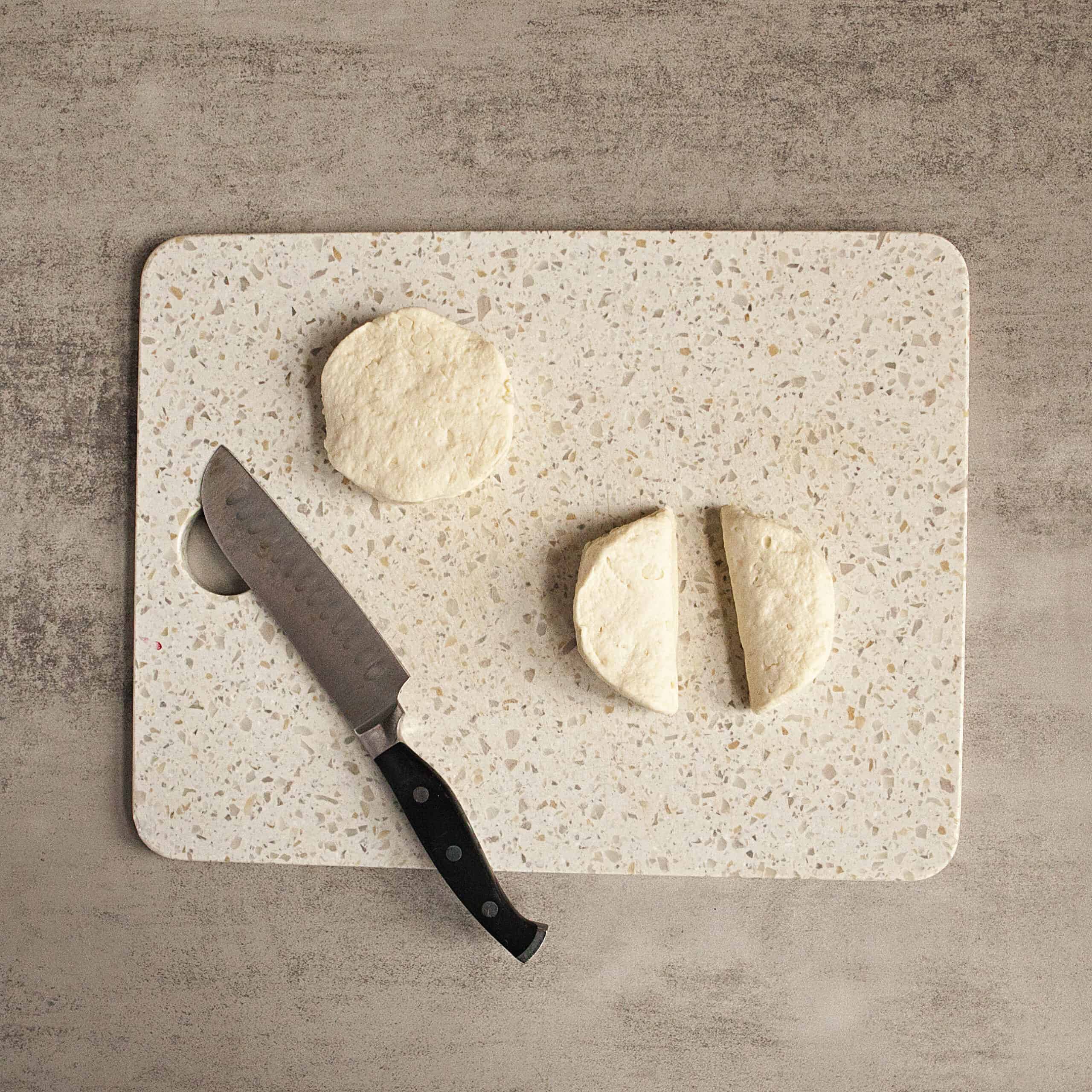 A white cutting board with a biscuit dough round and another biscuit dough round cut in half alongside a chef's knife.