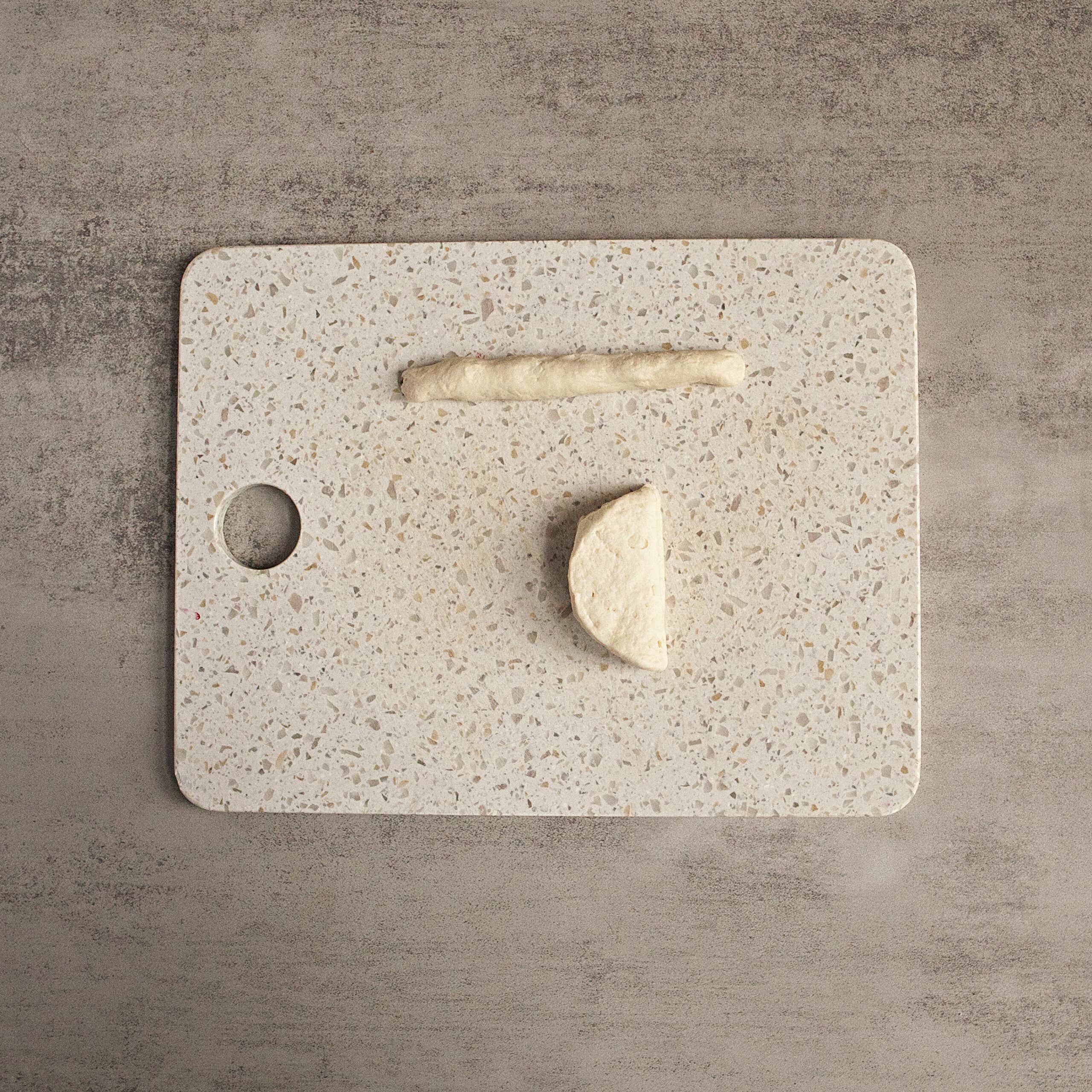 A white cutting board with half a round of biscuit dough and another piece of biscuit dough rolled into a 6-inch rope.