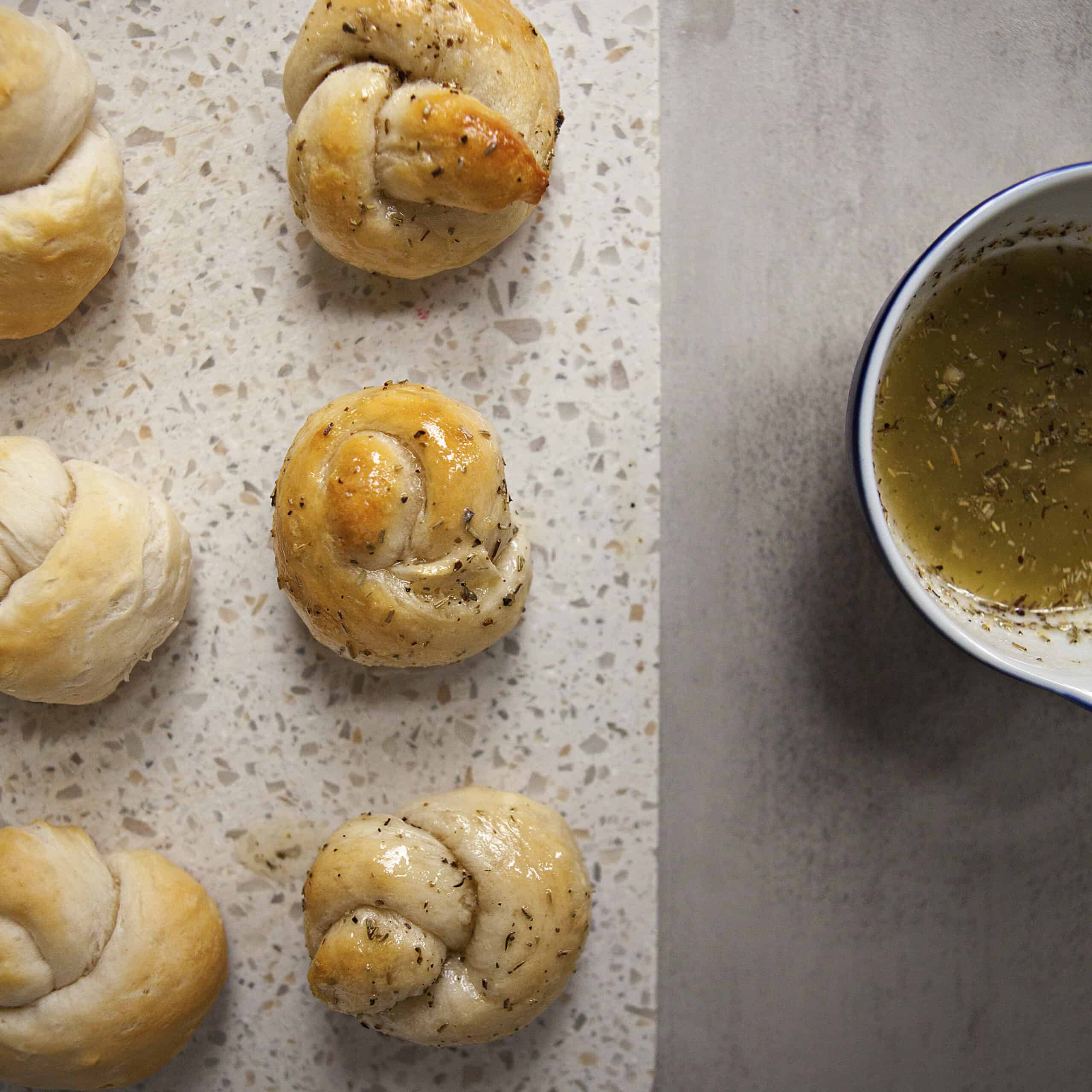 Garlic knots on a white cutting board next to a ramekin containing garlic butter.  Three of the knots have been dipped into the garlic butter.