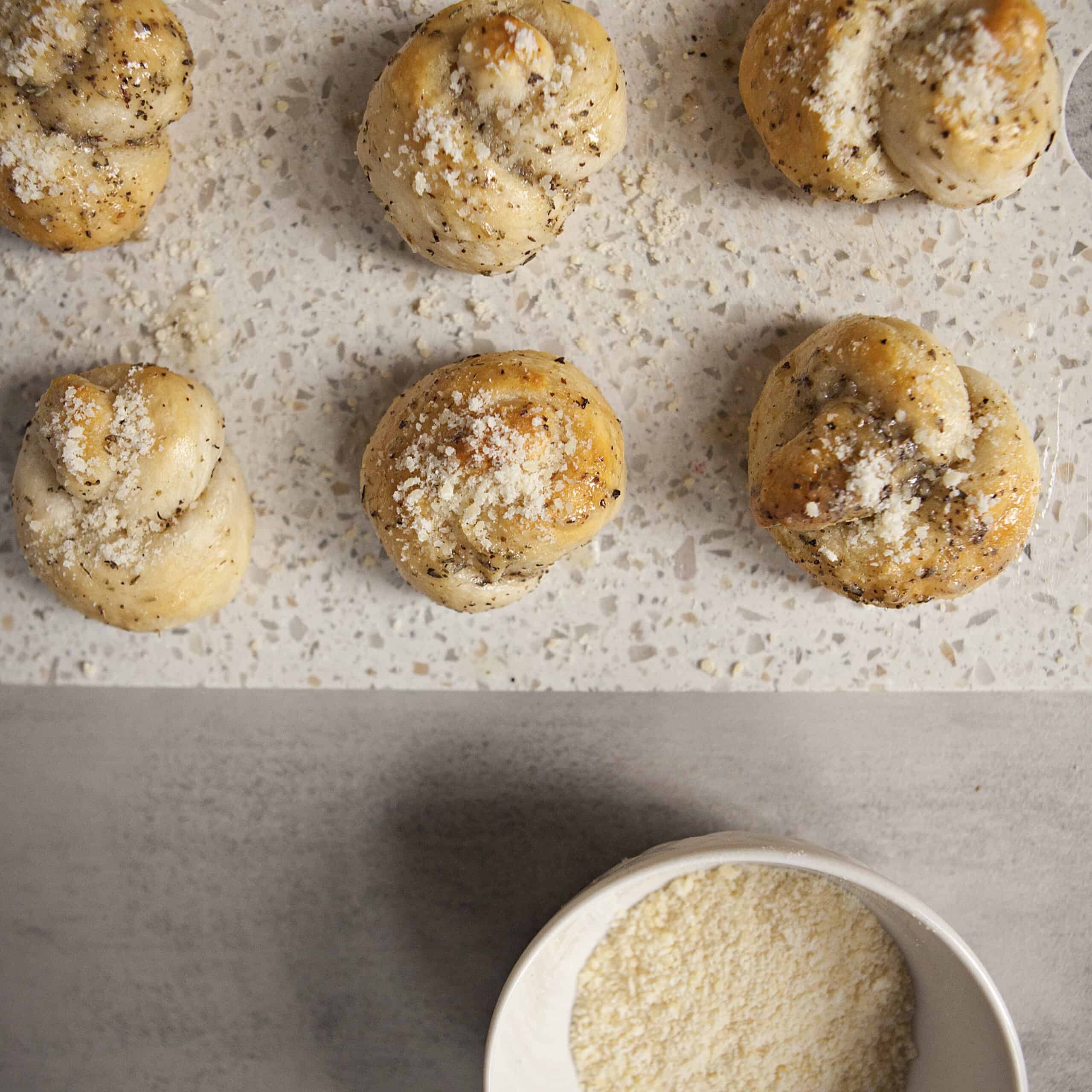 Buttery garlic knots on a white cutting board that have been sprinkled with grated Parmesan cheese, alongside a white ramekin containing more grated Parmesan cheese.