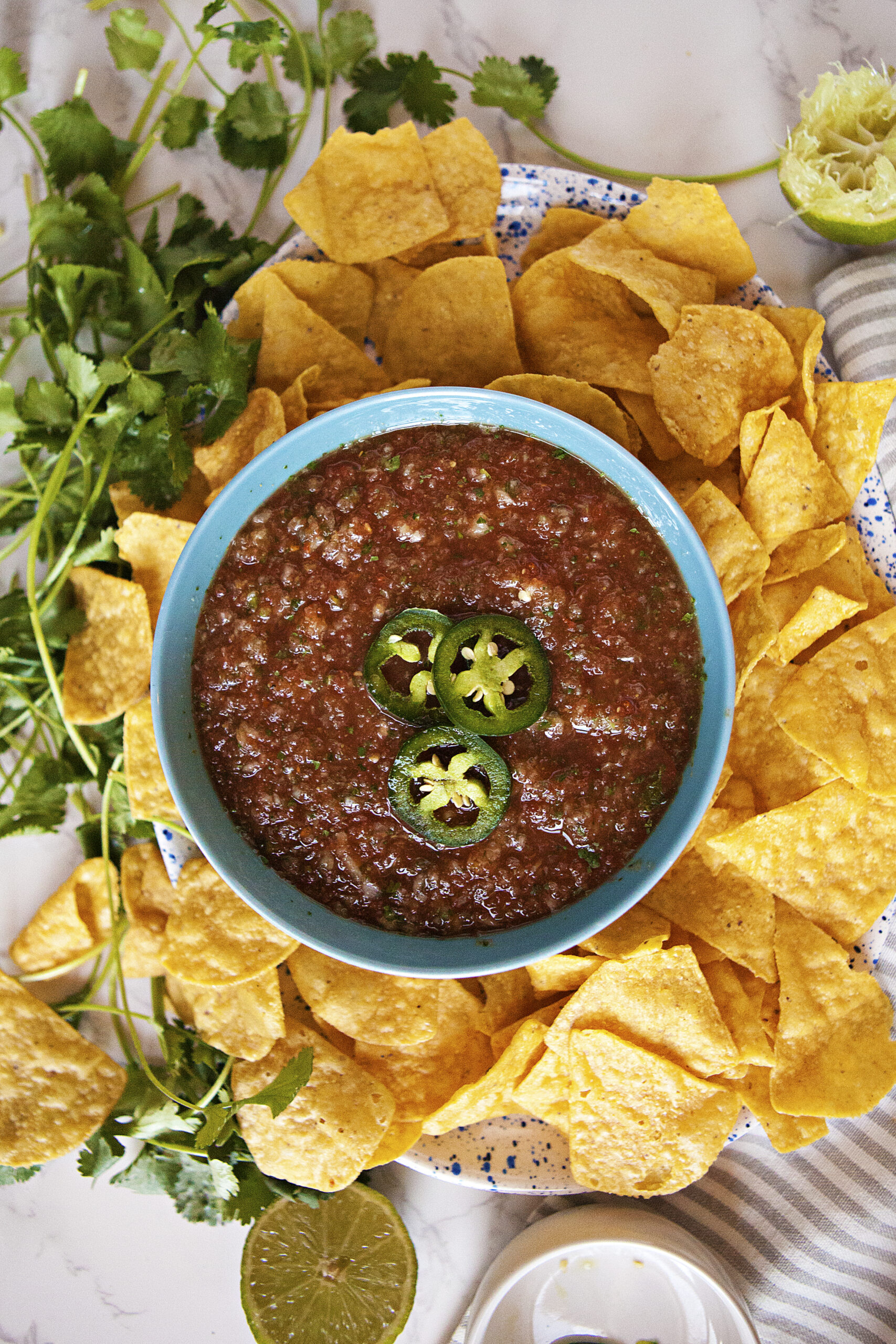 Restaurant-style salsa roja in a blue bowl topped with sliced jalapenos, alongside tortilla chips and fresh cilantro.