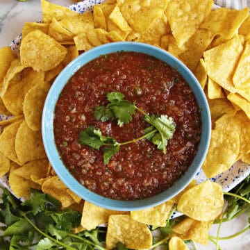 Restaurant-style blender salsa in a blue bowl, topped with fresh cilantros prigs, surrounded by tortilla chips.