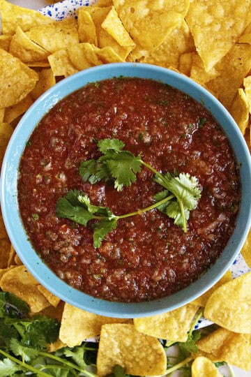 Restaurant-style blender salsa in a blue bowl, topped with fresh cilantros prigs, surrounded by tortilla chips.