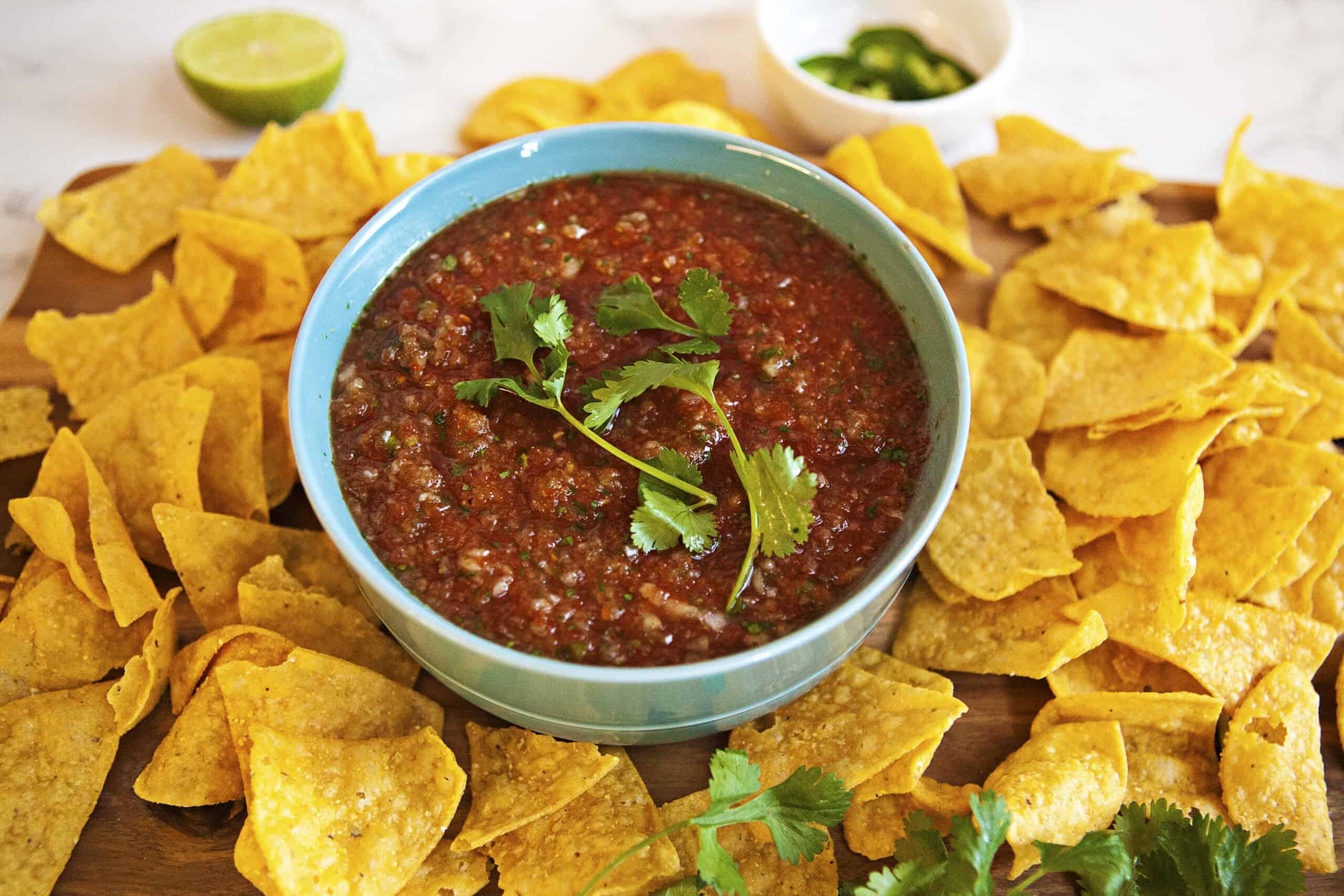 Restaurant-style blender salsa in a blue bowl, topped with fresh cilantros prigs, surrounded by tortilla chips.