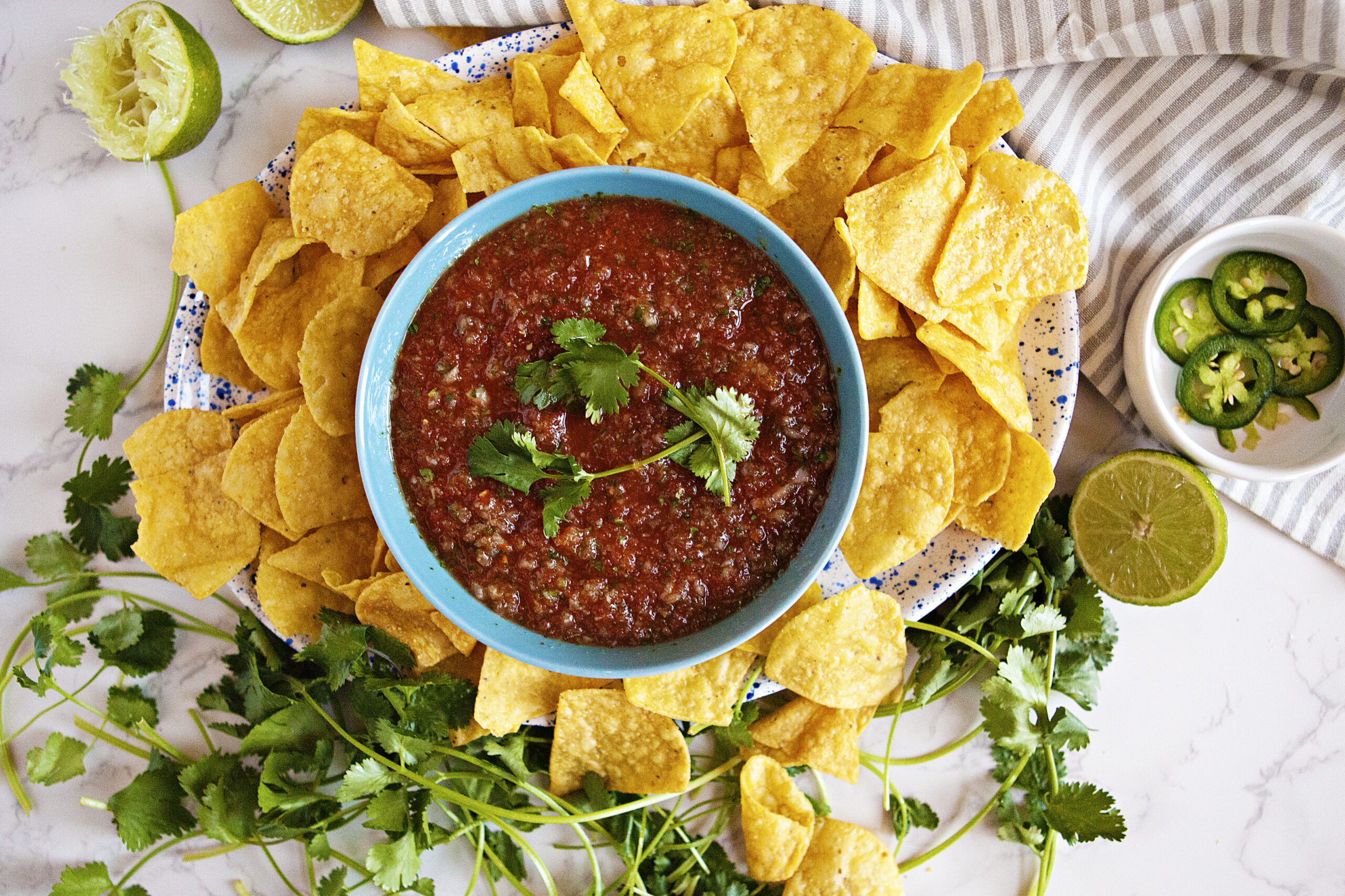 Restaurant-style homemade salsa in a blue bowl alongside tortilla chips, fresh cilantro, sliced lime, and jalapeno slices in a bowl.