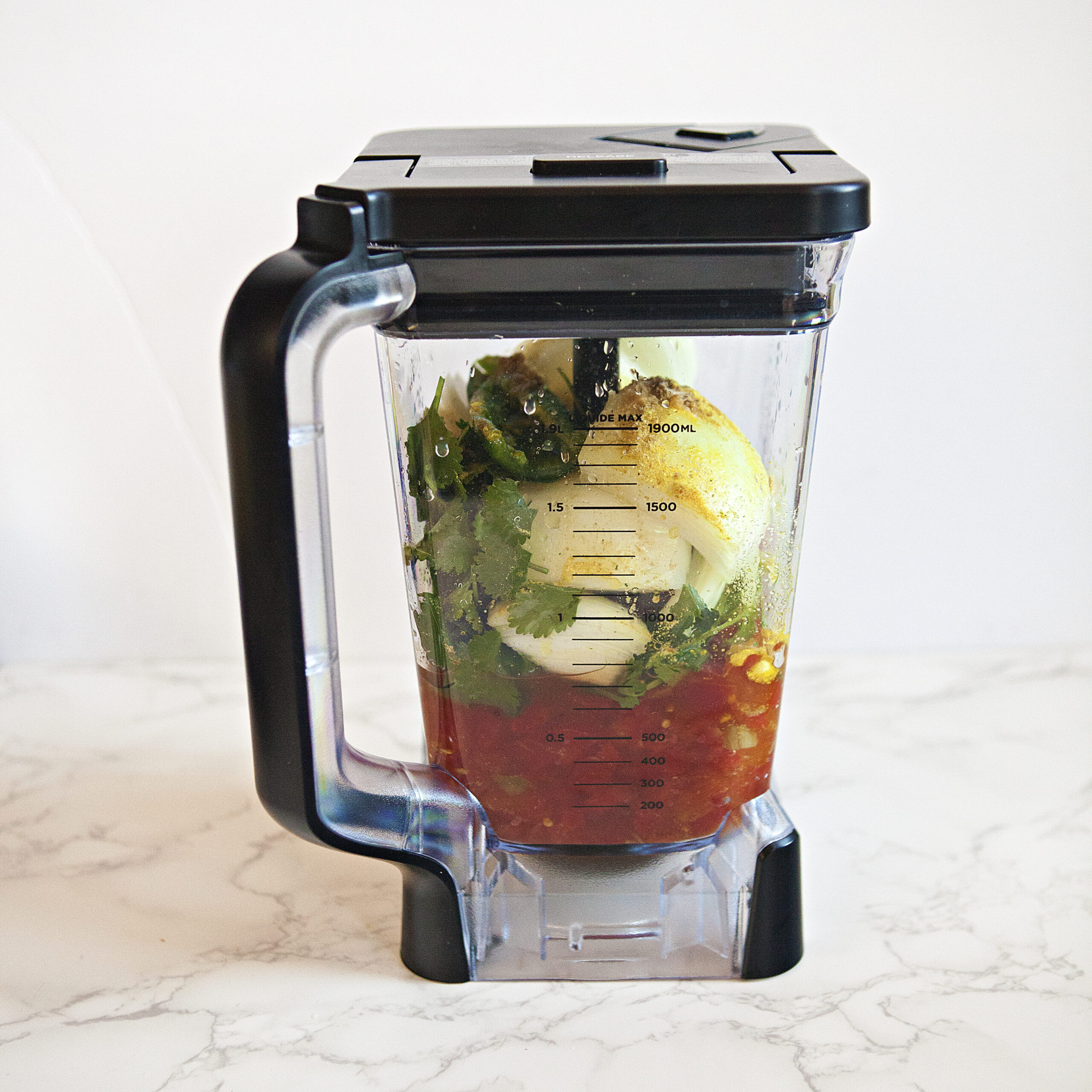 A blender containing all of the ingredients for restaurant-style salsa roja:  Tomatoes, green chilies, onion, cilantro, jalapeno, garlic powder, salt, cumin, chicken bouillon powder, and lime juice.