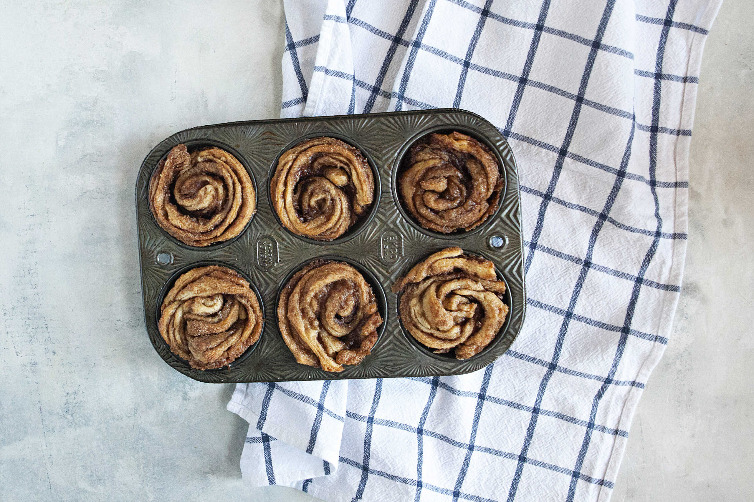 Cinnamon sugar cruffins in a muffin tin on top of a white and blue checked kitchen towel.