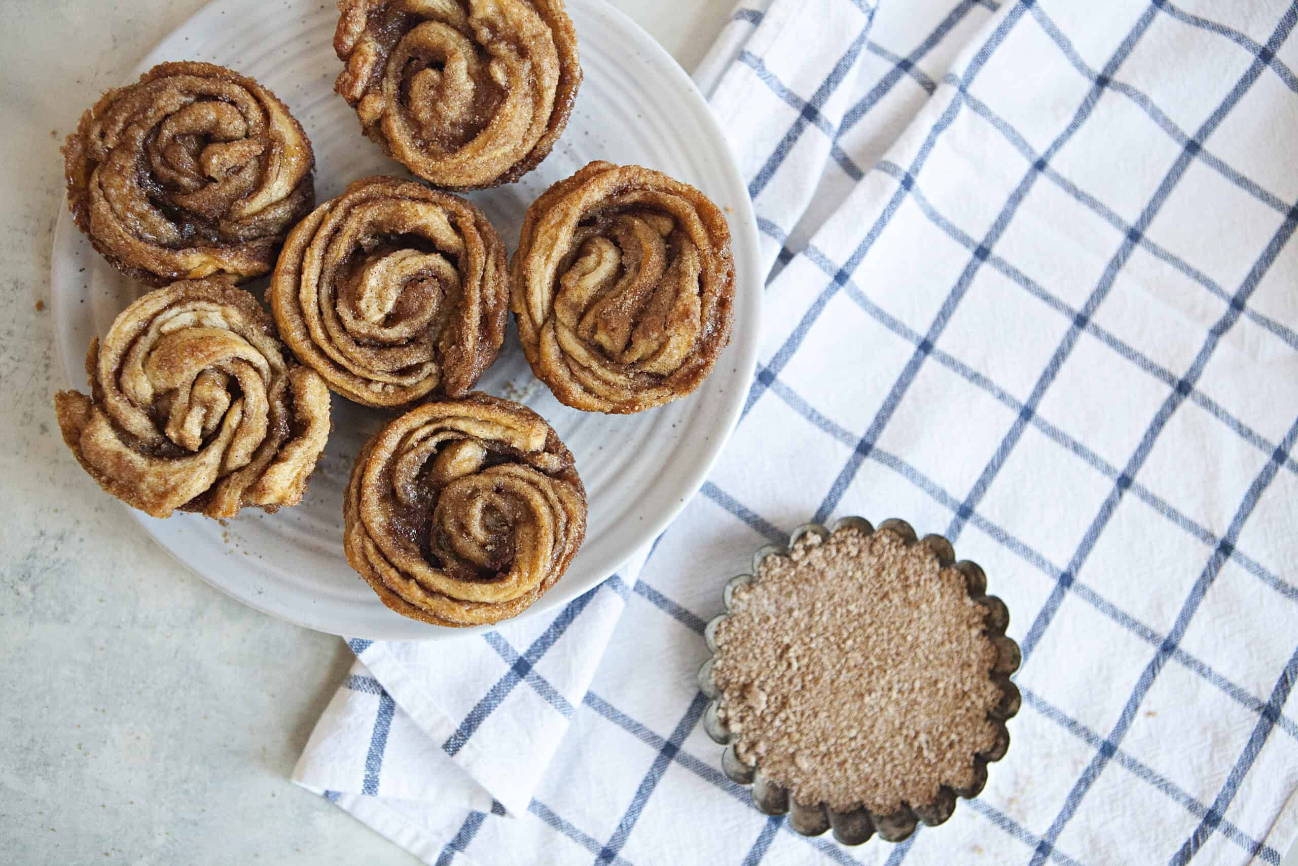 Cinnamon-sugar cruffins on a white plate alongside a small bowl of cinnamon-sugar and a white and blue checked kitchen towel.