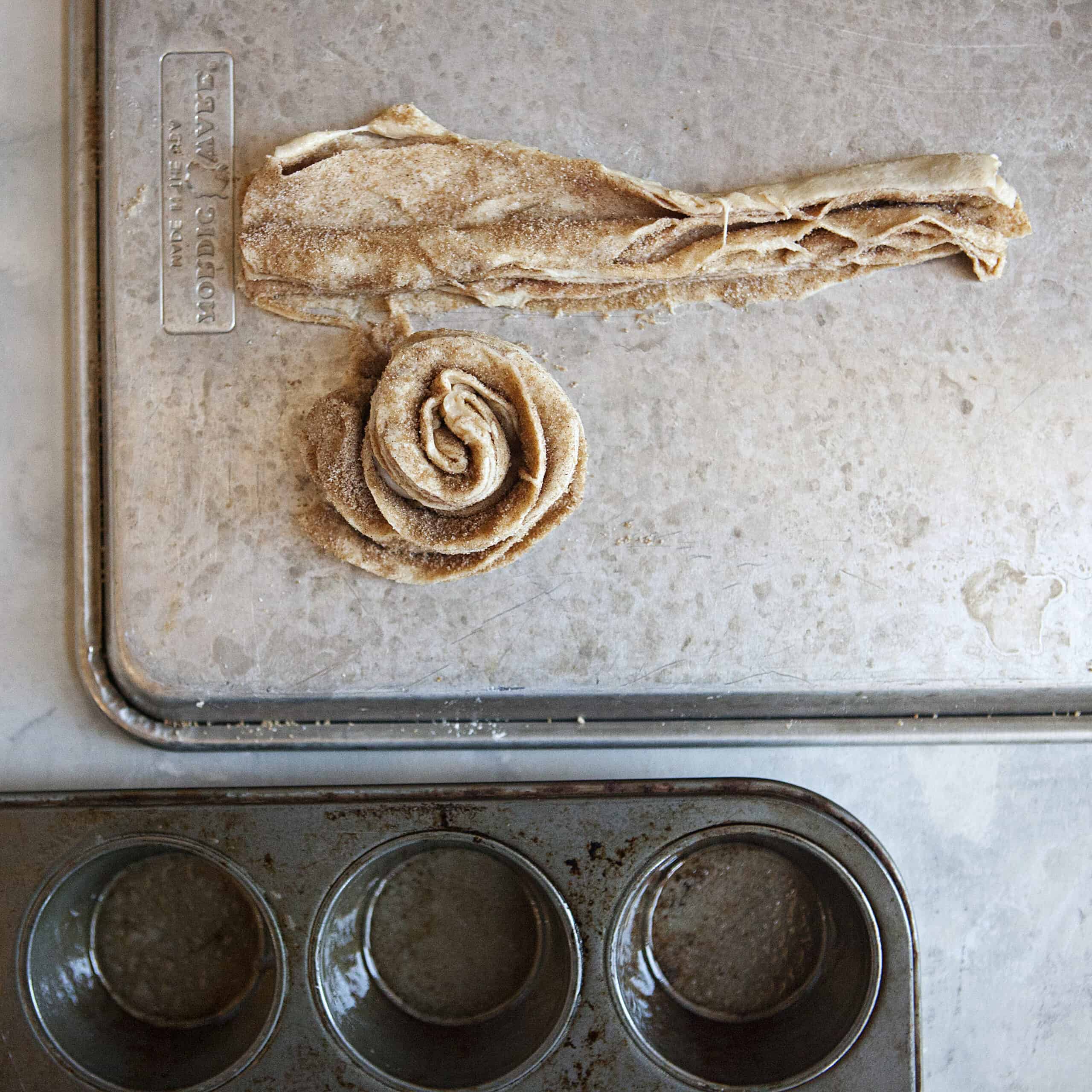 A log of crescent roll dough next to a unbaked cinnamon-sugar cruffin made of crescent roll dough and cinnamon-sugar on a metal baking sheet alongside a muffin tin.