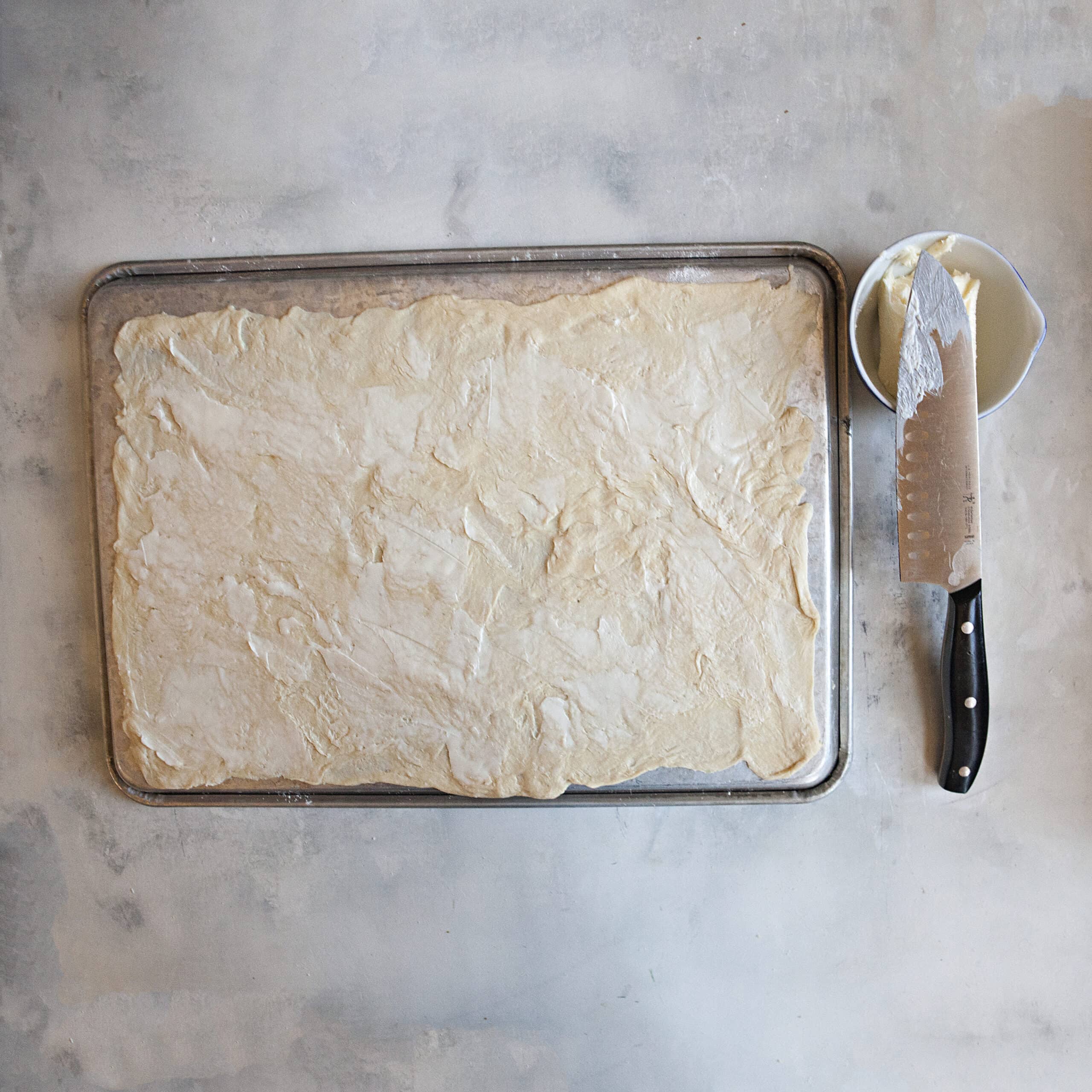 Lightly buttered crescent roll dough rectangle next to a small bowl with more butter and a chef's knife.