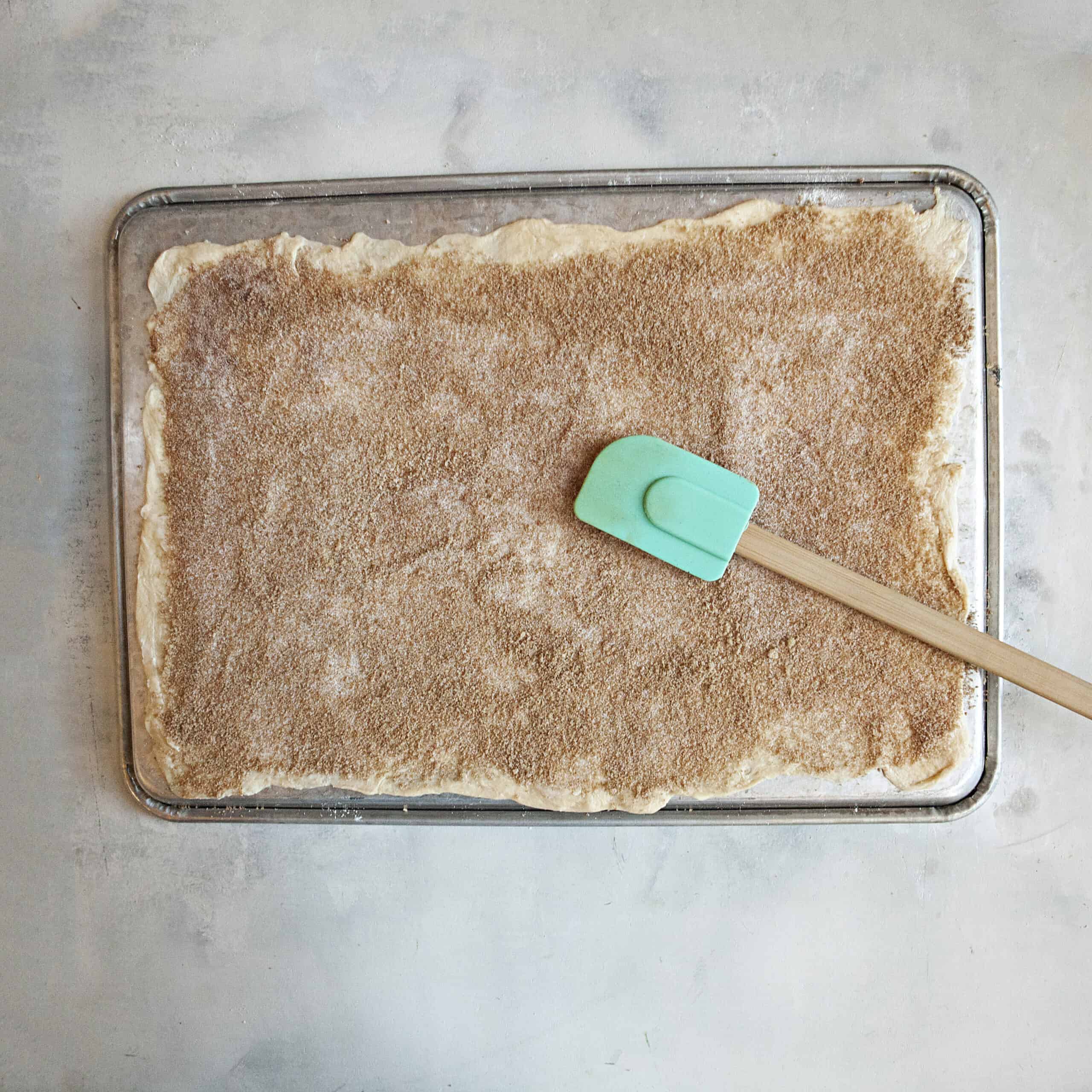 A rectangle of crescent roll dough topped with cinnamon-sugar and a turquoise spatula.