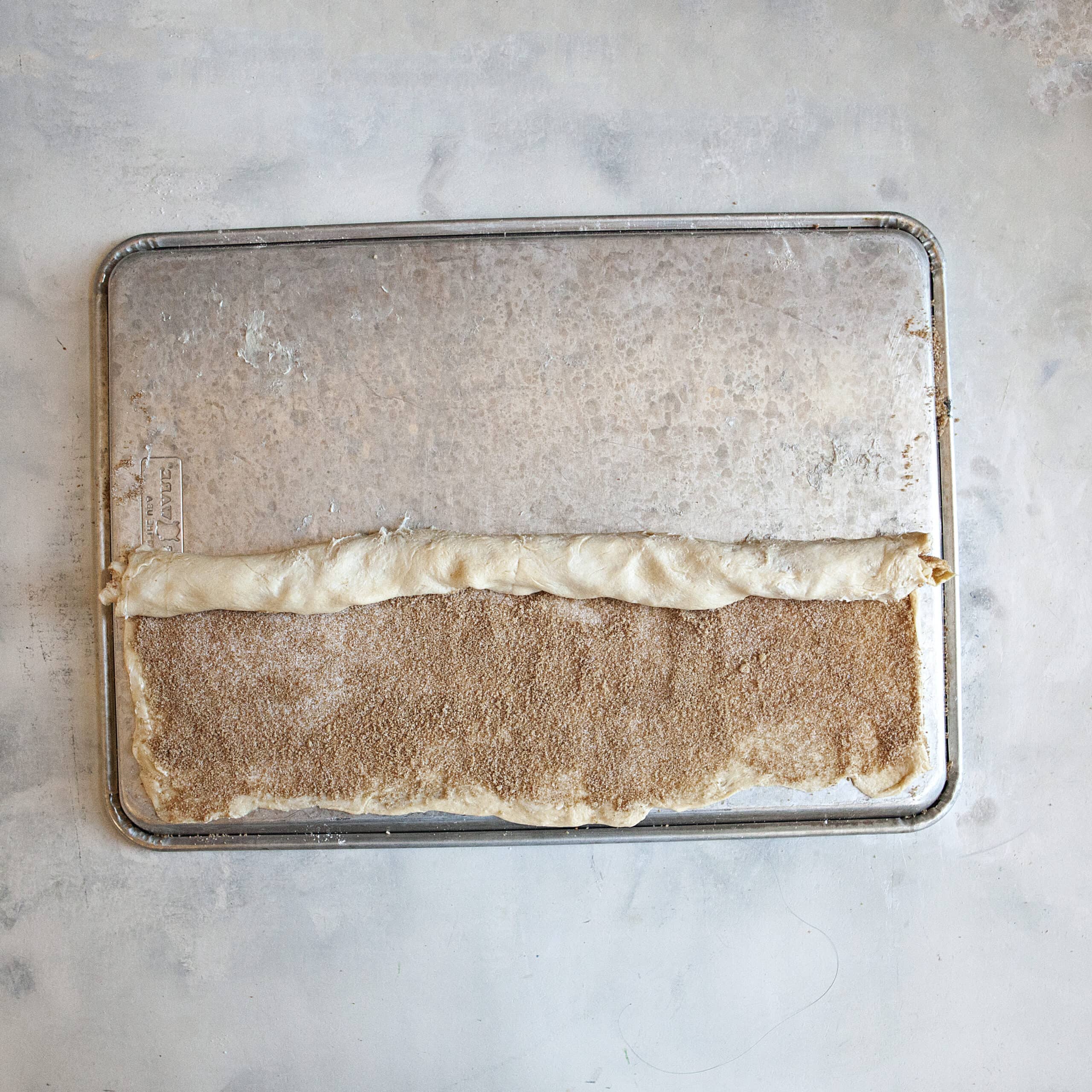 A rectangle of crescent roll dough covered with cinnamon-sugar being rolled up lengthwise on top of a metal baking sheet.