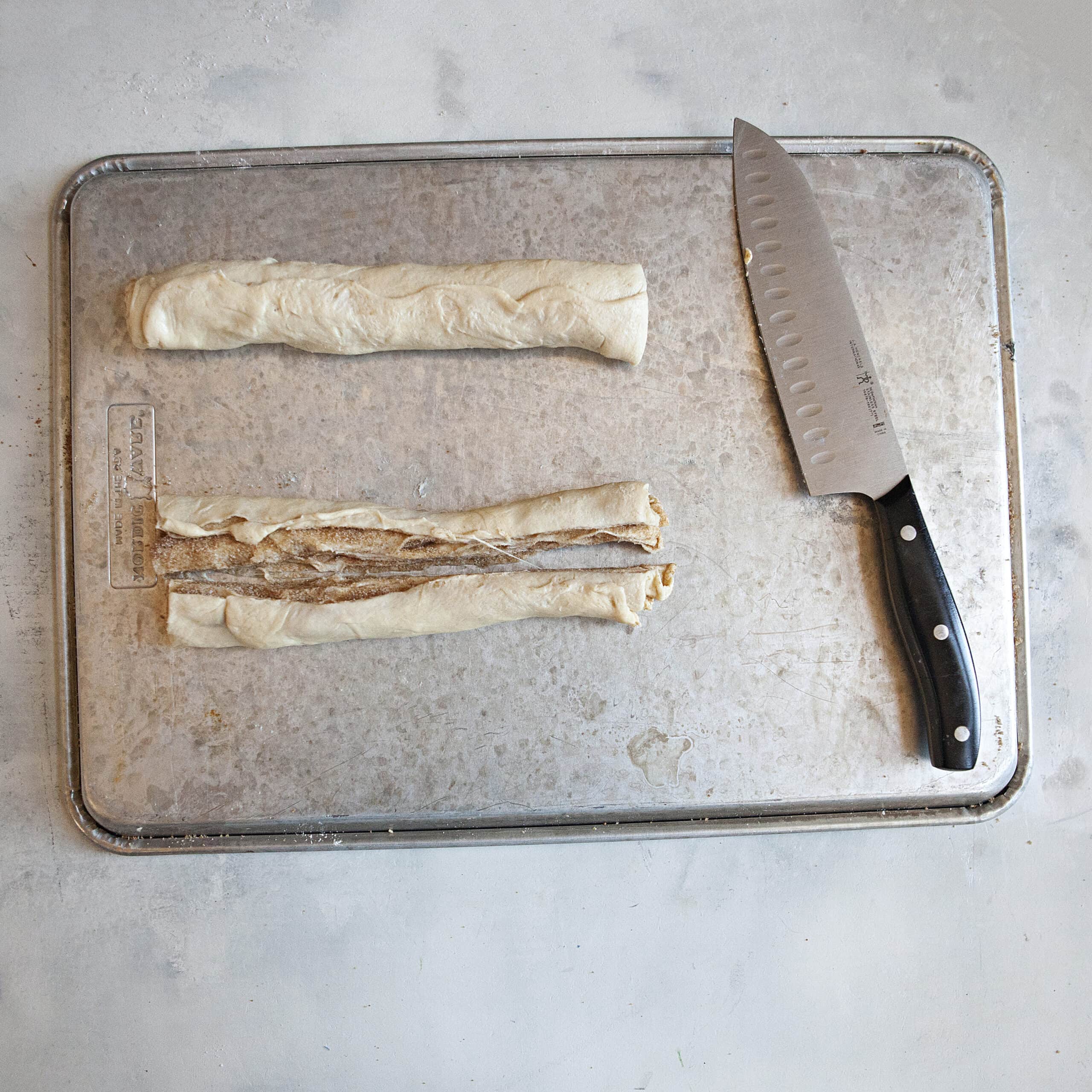 A cylinder of crescent roll dough alongside another roll of crescent roll dough cut in half lengthwise as well as a chef's knife on a metal baking sheet.