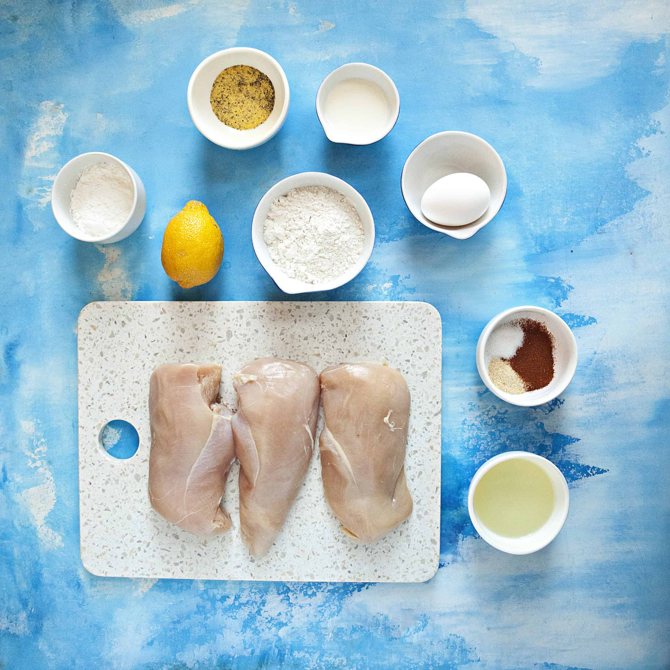 Ingredients for lemon pepper chicken bites:  Chicken breasts, flour, cornstarch, lemon pepper seasoning, garlic powder, paprika, salt, egg, milk, lemon juice, and oil, on a blue backdrop.