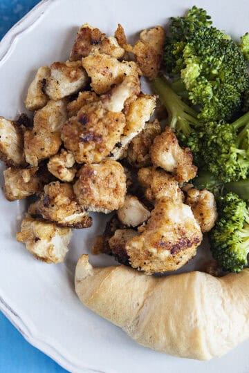 Lemon pepper chicken bites on a white plate with broccoli and a roll.