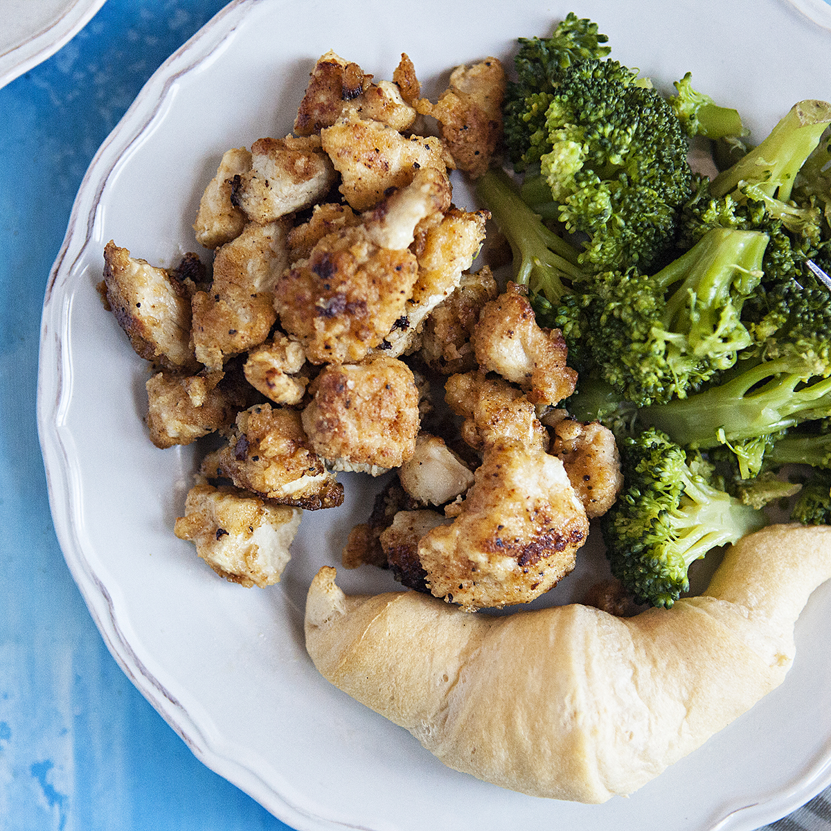 Lemon pepper chicken bites on a white plate with broccoli and a roll.