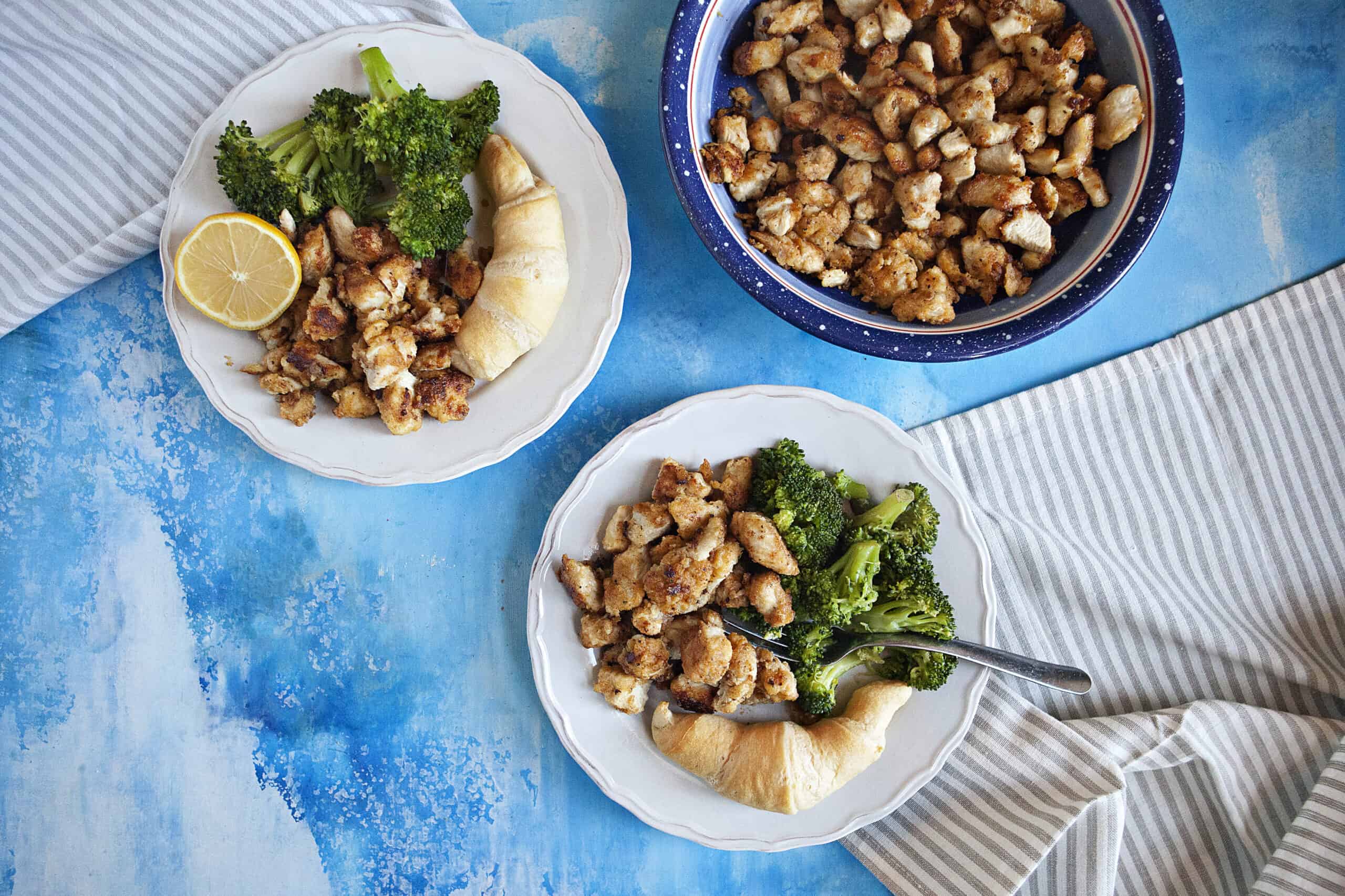 Two plates of lemon pepper chicken bites, broccoli, and rolls with a fork alongside a gray and white striped napkin and another bowl containing more lemon pepper chicken, all on a blue backdrop.
