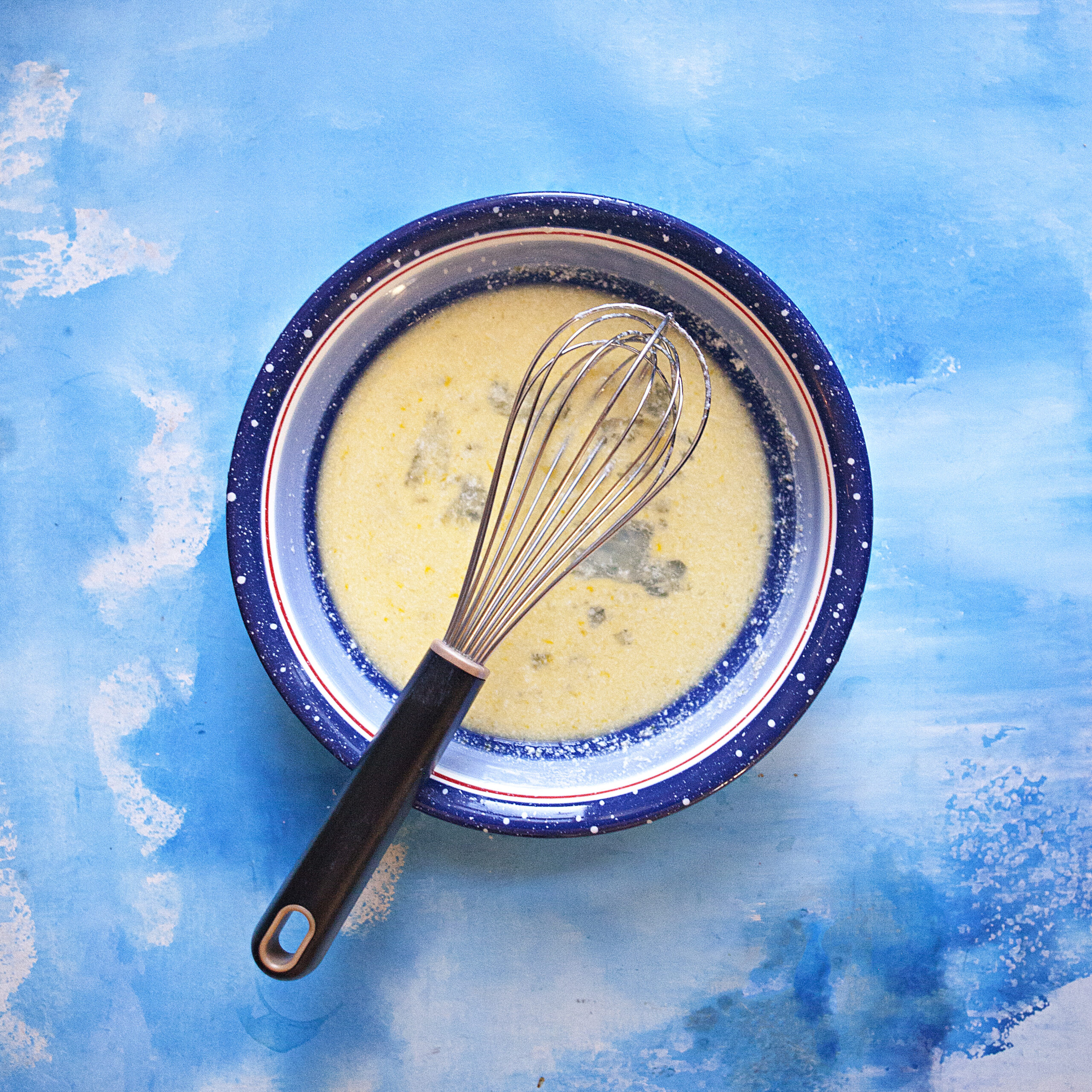 Egg wash made of egg, milk, and lemon juice in a blue bowl with a whisk, on top of a blue backdrop.